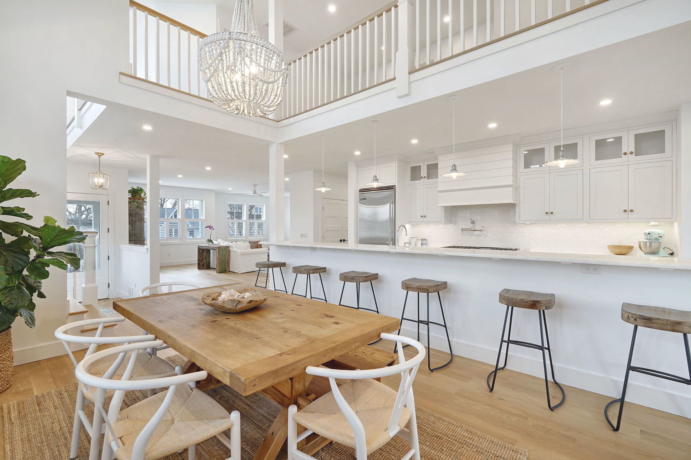 Open-concept kitchen and dining area with a high ceiling, white cabinets, a stainless steel refrigerator, and a wooden dining table surrounded by white chairs. A large chandelier hangs from the ceiling, and there is a green plant near the entrance.