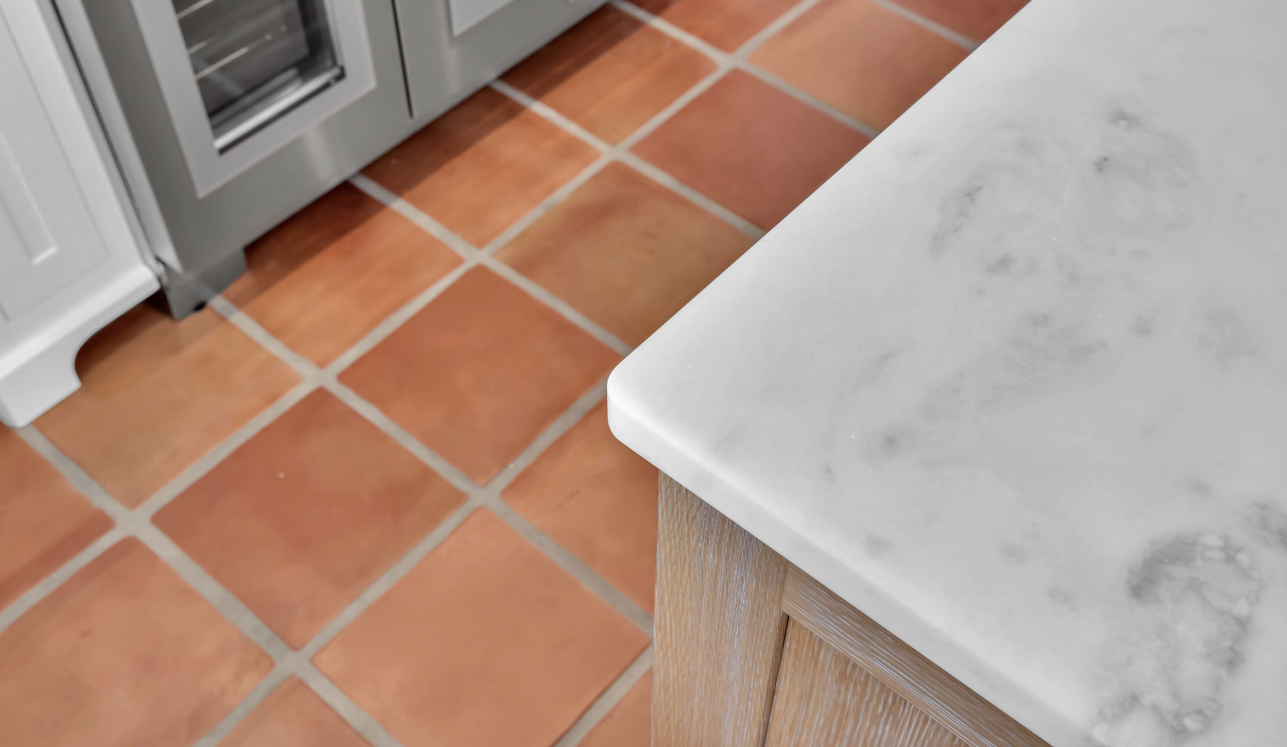 Close-up of a kitchen countertop with a marble surface and a wooden cabinet corner, with terra cotta tiled flooring in the background.