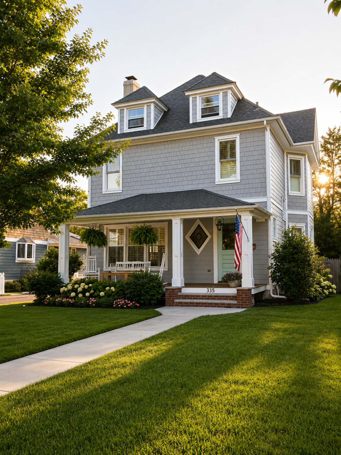 A three-story gray house with white trim, a porch with hanging plants, an American flag, and a front yard with green grass and flowers.