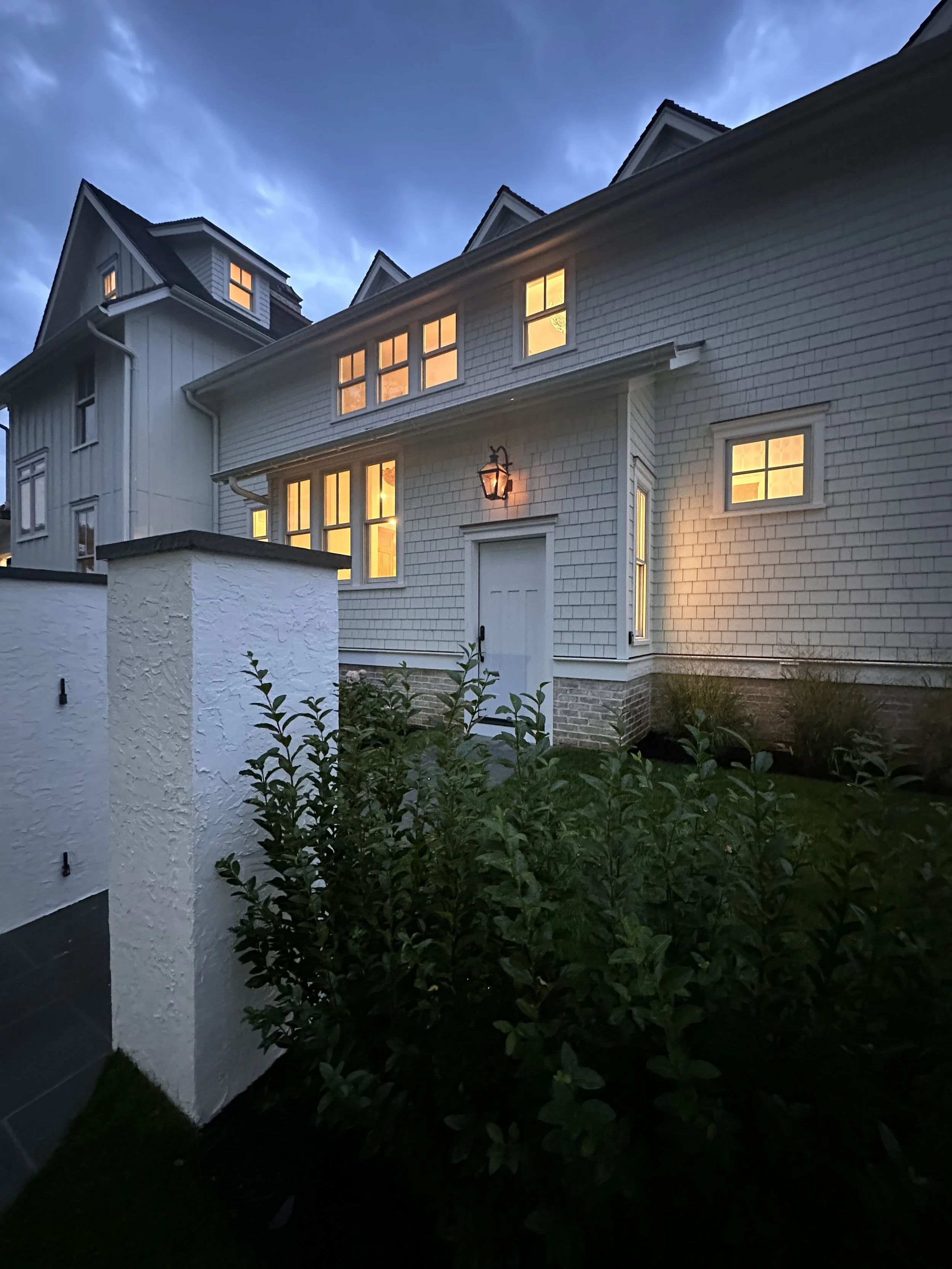 A white house with multiple windows glowing with yellow light at dusk. There are darkening blue skies with some clouds and a small garden with leafy bushes in the foreground.