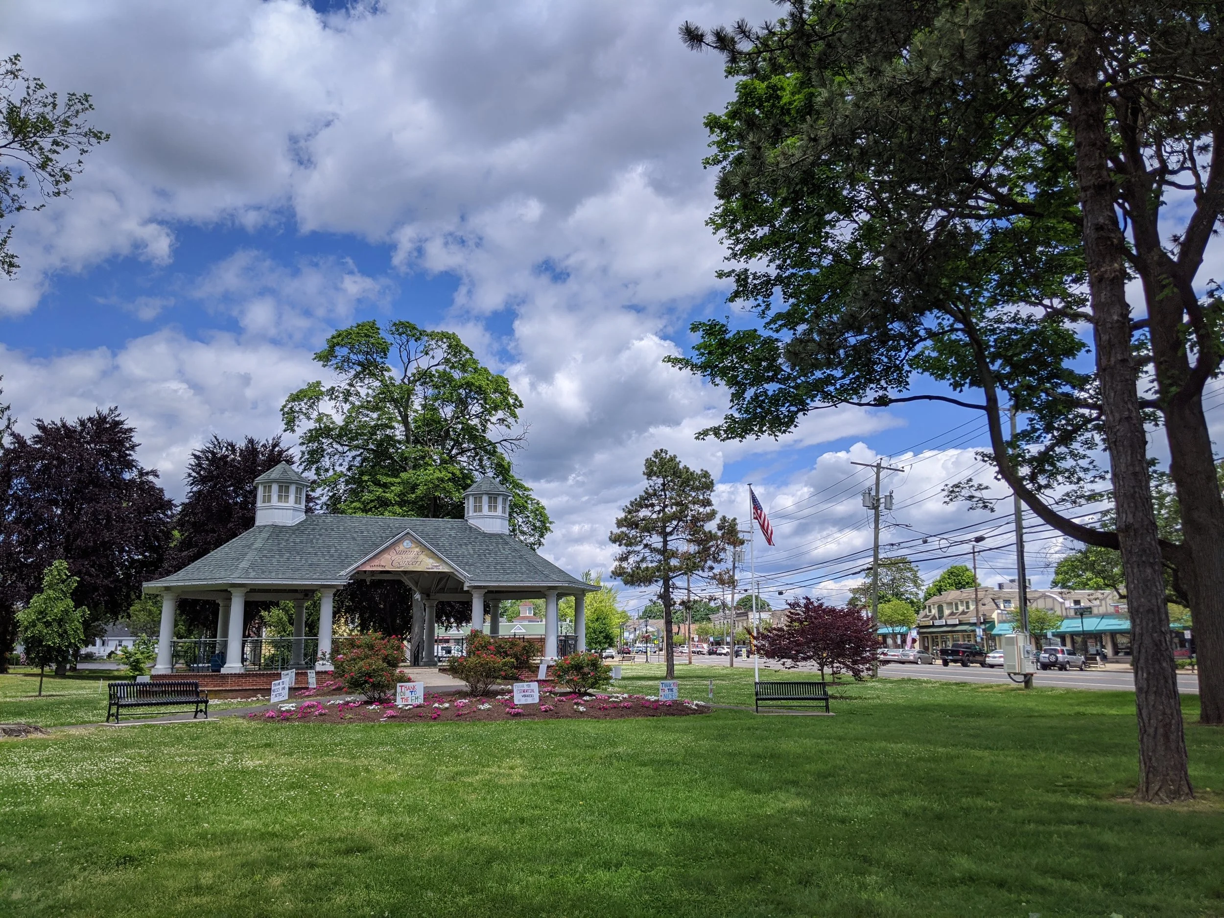 A park scene with a historic gazebo, surrounded by blooming flowers and green grass, with trees and a cloudy sky overhead.