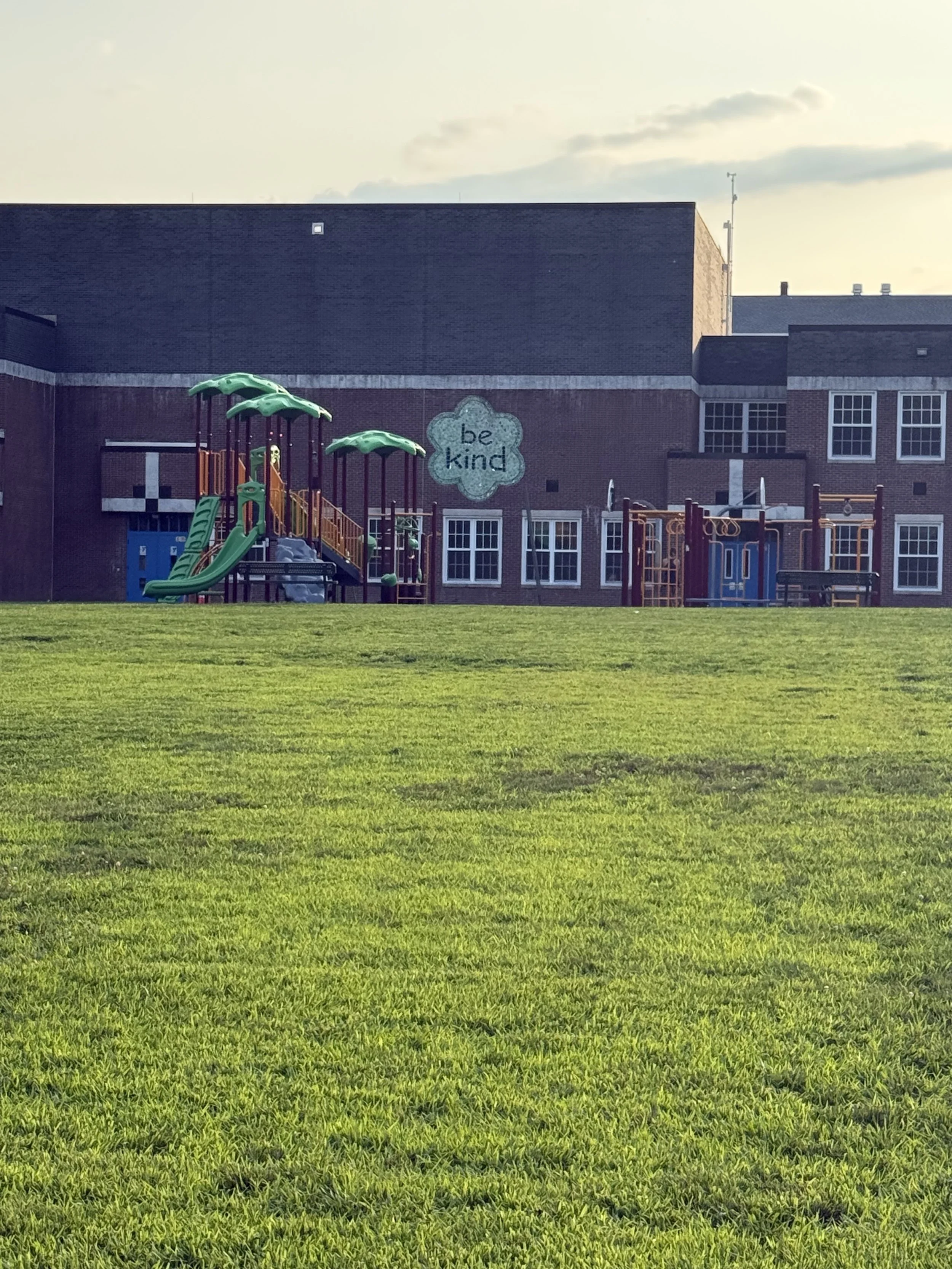A playground with a green slide and climbing structure in front of a brick school building. The building has multiple windows and a sign that says 'be kind'. The grassy field in the foreground is well-maintained, and the sky has some clouds with a hint of sunset light.