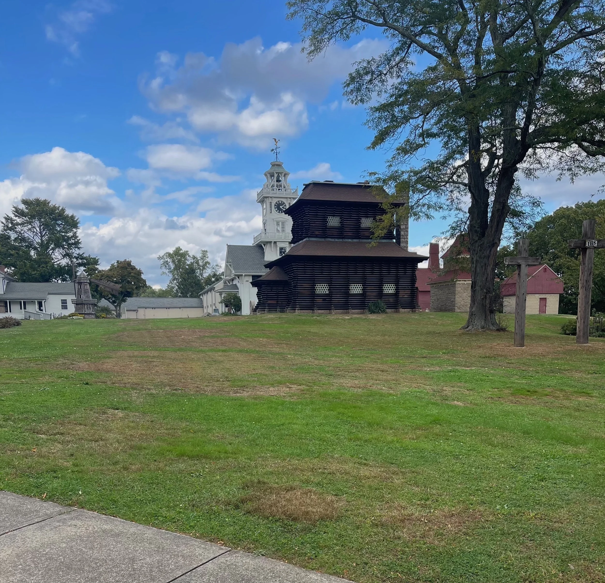 Lawn with green grass, a sidewalk in the foreground, a tree with green leaves on the right, and a historic white clock tower building with a weather vane on top in the background under a partly cloudy blue sky.