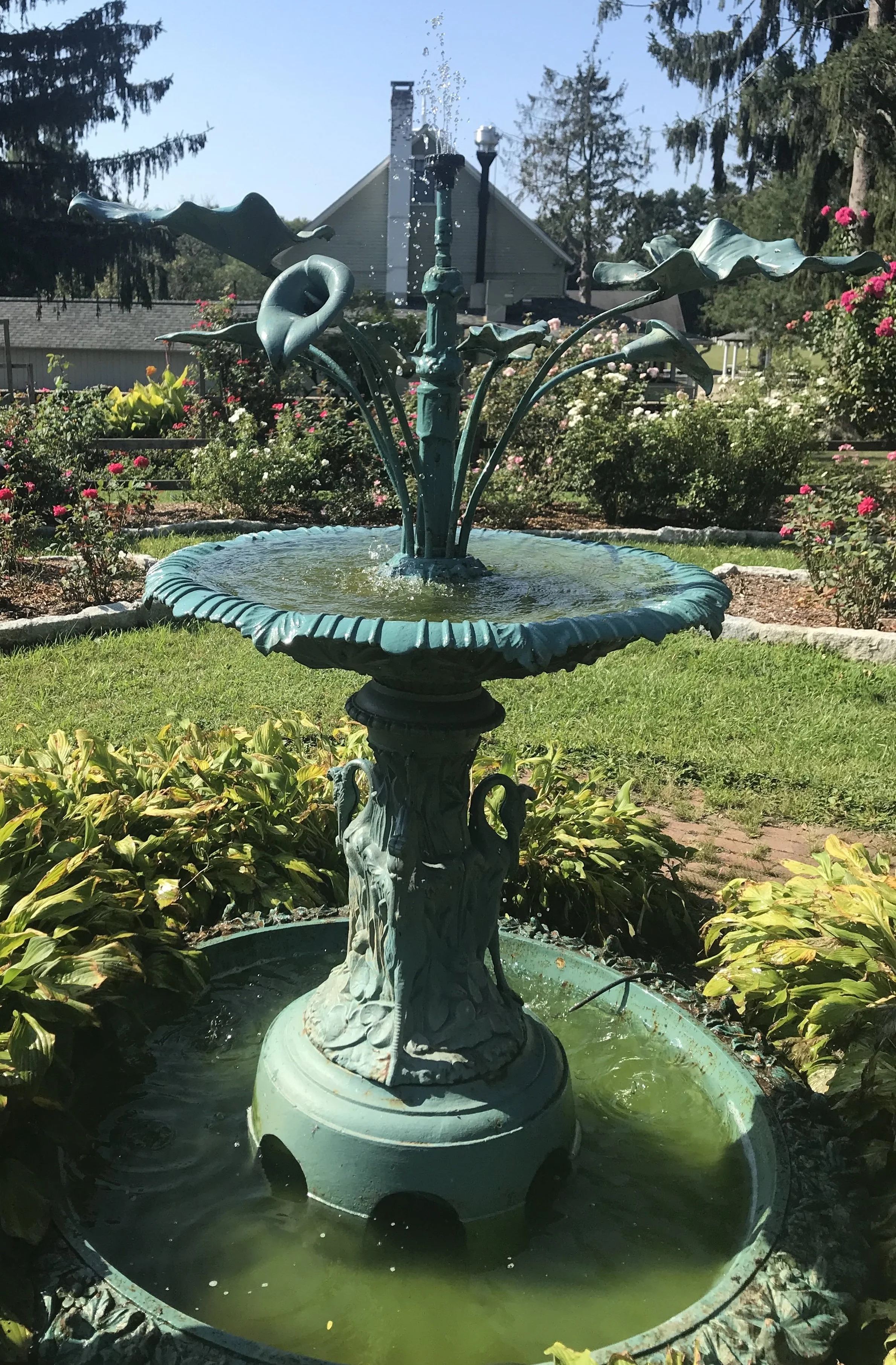 A decorative metal garden fountain shaped like a lily pad with a central pole adorned with leaves, surrounded by a pond filled with green water, in a flower garden with bushes, flowers, and trees in the background.