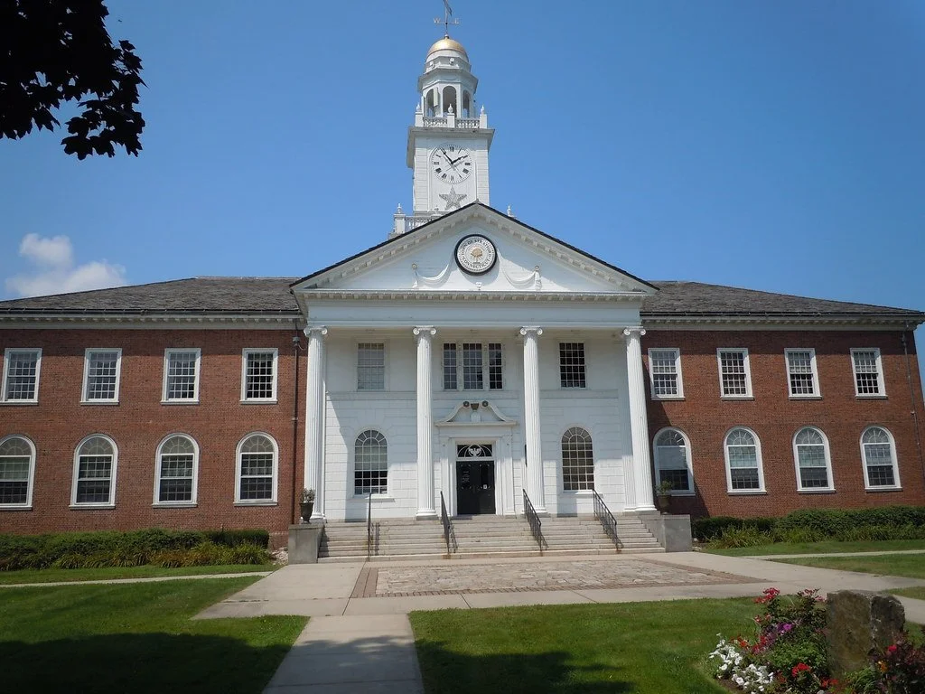 A historic white and red brick town hall with a prominent clock tower in the center, surrounded by a green lawn and flowers under a clear blue sky.