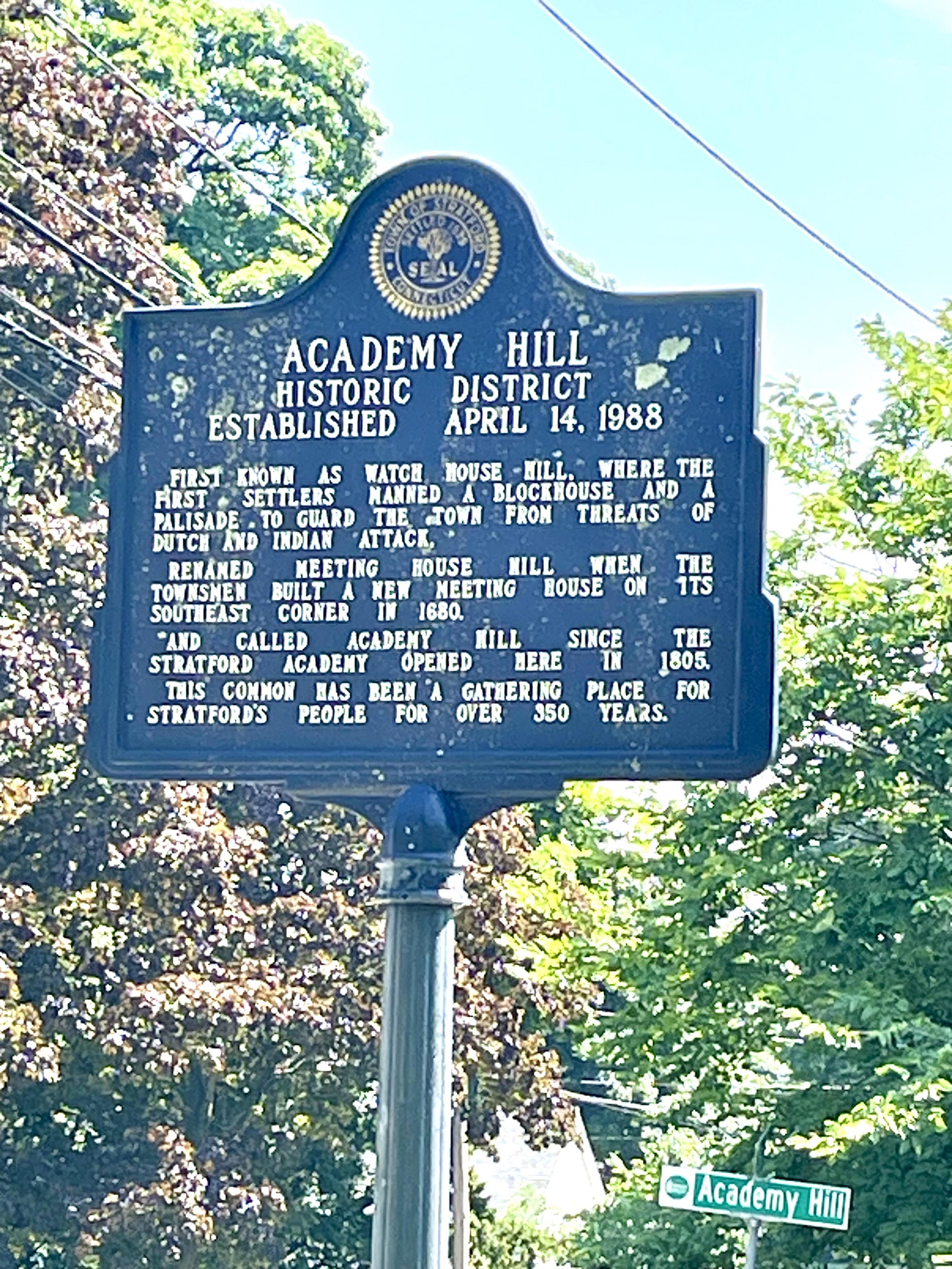 A historic blue street sign reads 'Academy Hill Historic District' established on April 14, 1988, with a circular seal at the top. A green street sign at the bottom right reads 'Academy Hill', with trees and power lines in the background.