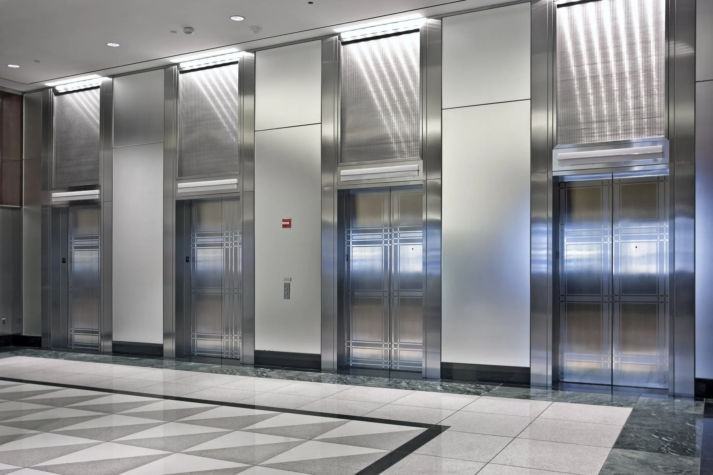 An elevator bank of four elevators within the interior of a modern building. The elevators are sleek, and of silver color.