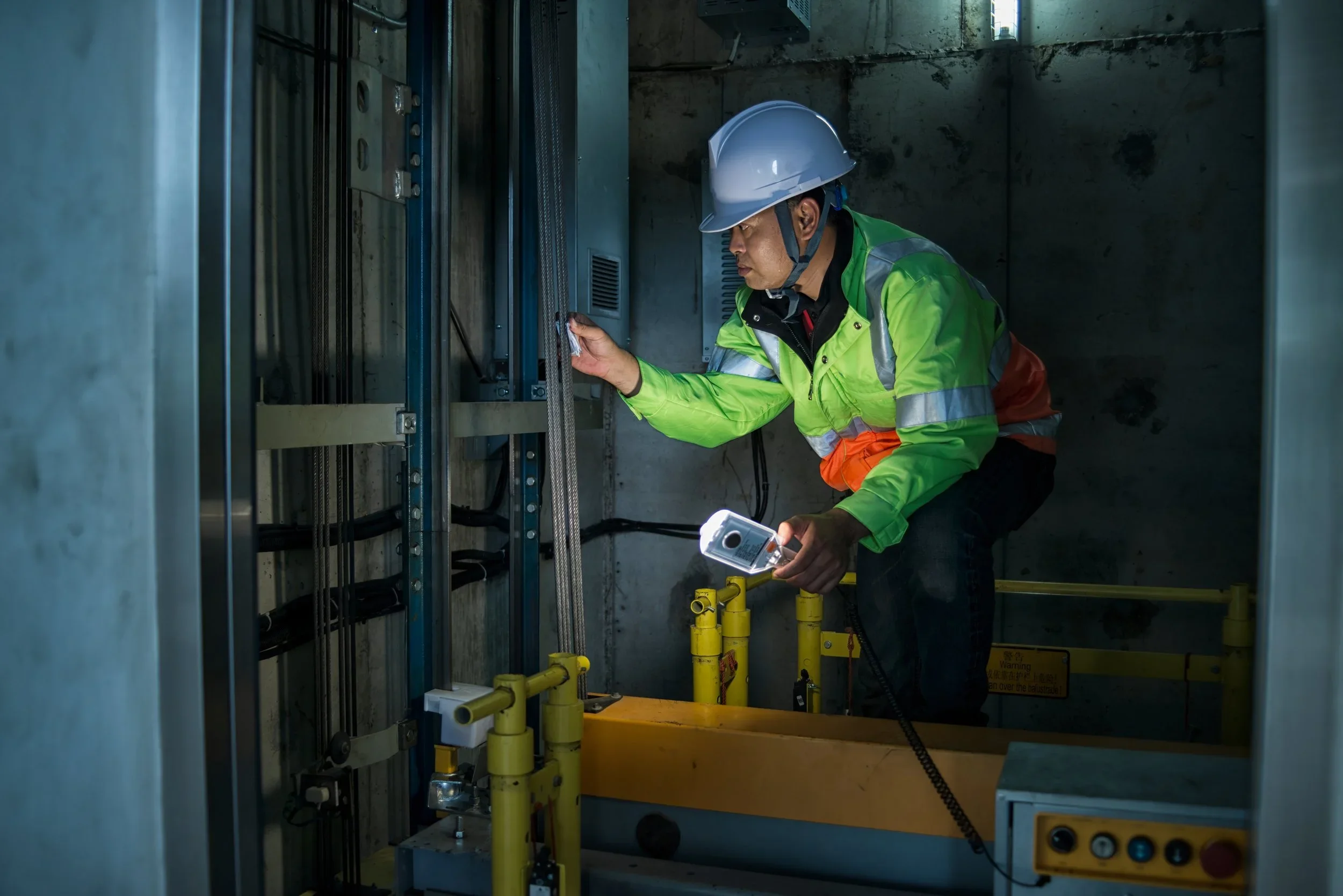 Image of a man in a florescent green jacket and hard hat, inspecting and standing within an open elevator shaft.