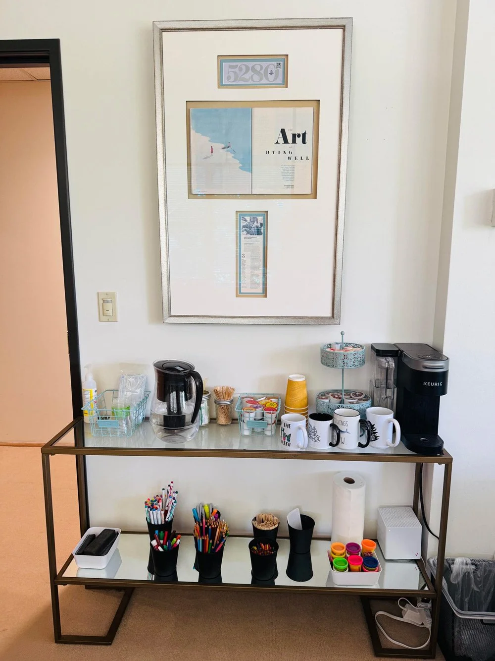 A refreshment station with a coffee maker, Keurig machine, mugs, cups, and an assortment of sweeteners and accessories on a two-tier glass table against a white wall with framed art.
