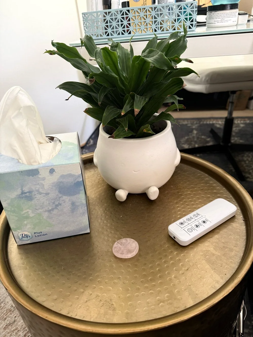 A potted green plant in a white ceramic pot on a round metallic gold table. Next to the plant, there is a box of tissues, a small round pink object, and a white remote control. In the background, there are shelves with various bottles and containers.