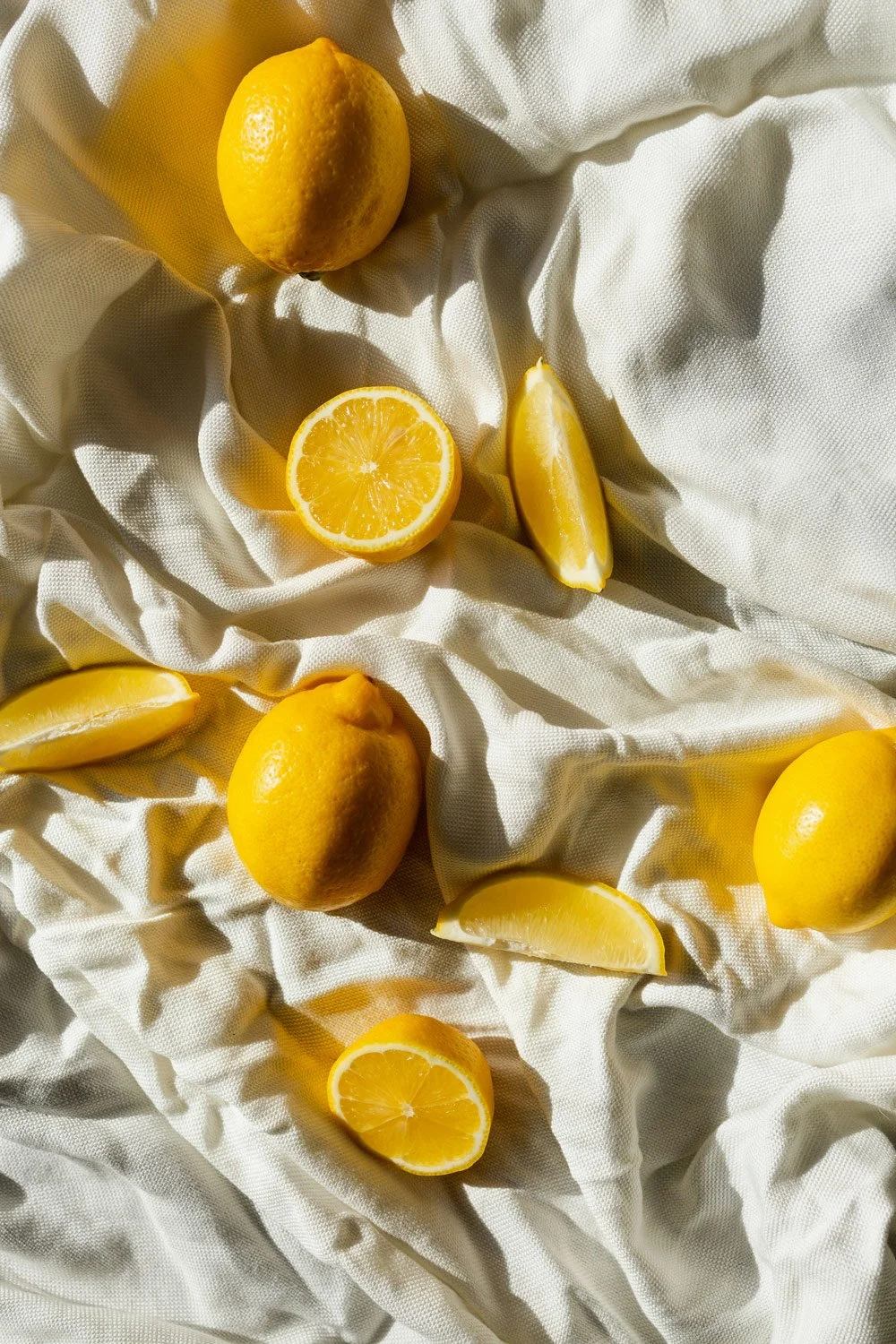 Lemons, some whole, some cut in half or wedges, arranged on crumpled light-colored fabric.