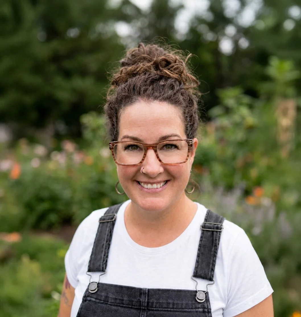 Hannah Dudley, LMFT is a smiling woman outdoors with glasses, curly hair in a bun, wearing a white t-shirt and black overalls.