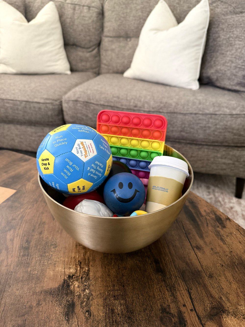 A silver bowl filled with various colorful toys and objects on a wooden table in front of a gray sofa with white pillows.