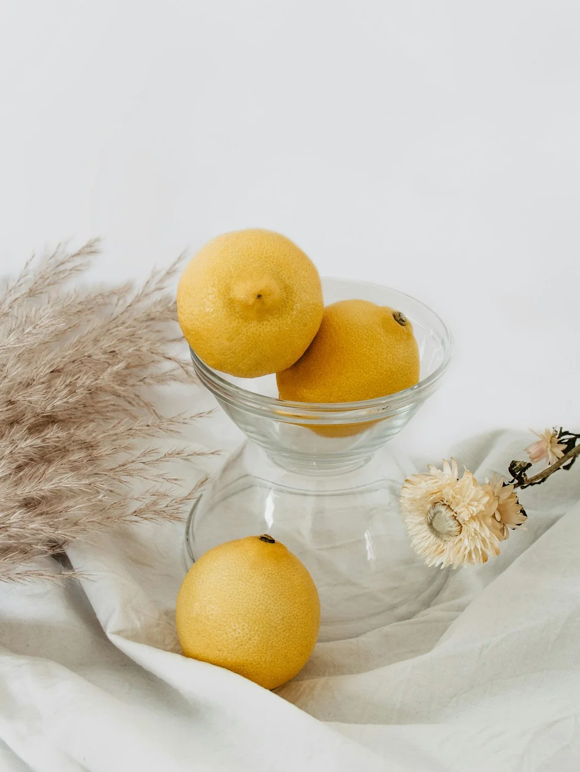 Three yellow lemons in a glass bowl, with one lemon outside the bowl, next to a dried flower and pampas grass on a white cloth background.