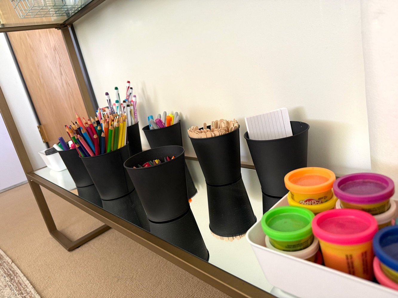 Organized black containers on a table holding colored pencils, markers, crayons, and Play-Doh containers.