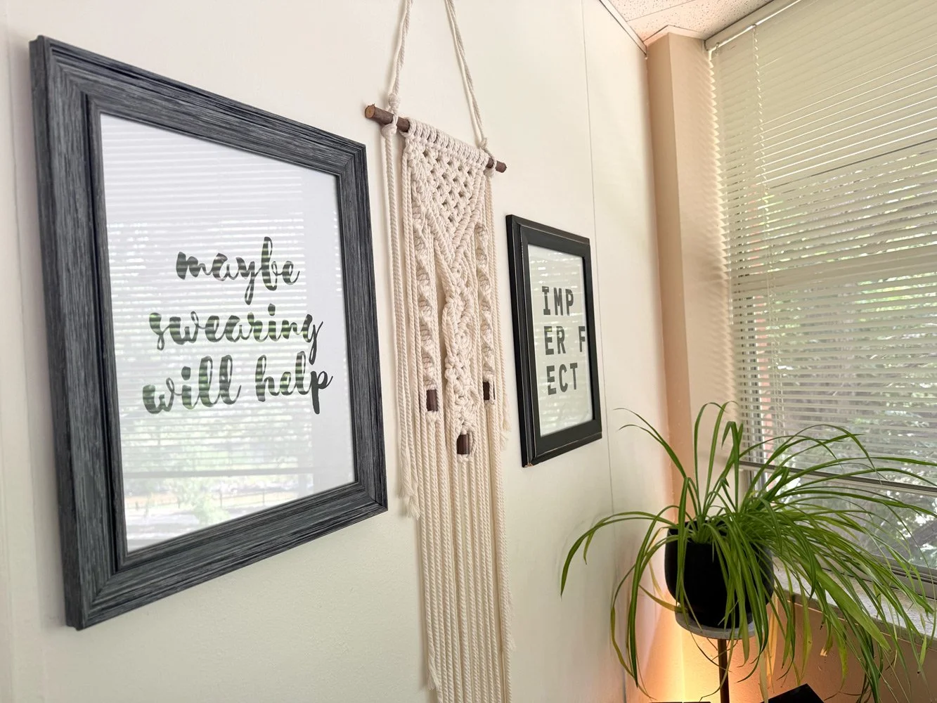 Decorative wall with framed signs, a macramé wall hanging, and a potted plant beside a window with blinds.