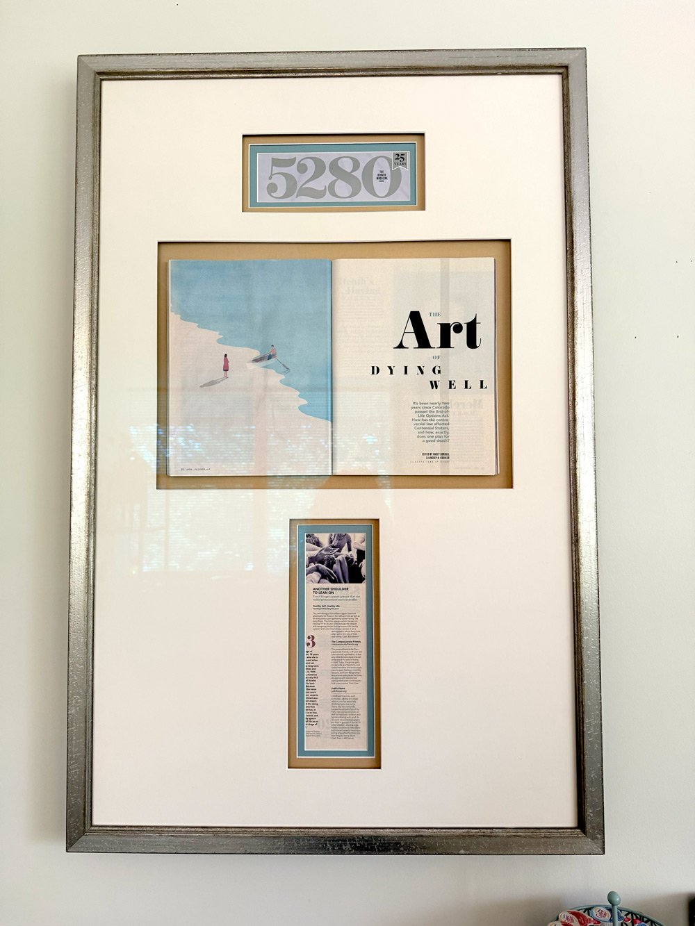 Framed display with a large number '5280' at the top, an open magazine with a beach scene and the title 'The Art of Dying Well,' and a vertical list with an image at the bottom.