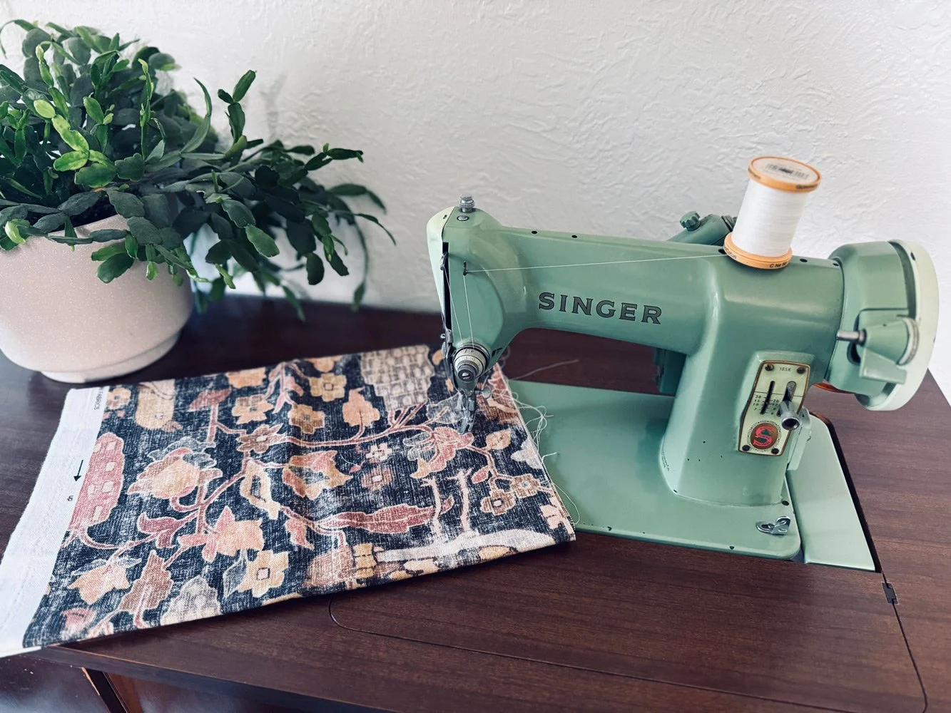 A vintage green Singer sewing machine on a wooden table with a partially sewn floral fabric, next to a potted green plant against a white textured wall.