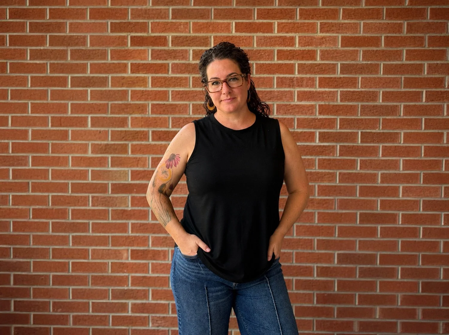 Trauma and grief therapist Hannah Dudley, LMFT is a woman with dark, curly hair, glasses, and hoop earrings stands in front of a red brick wall with tattoos on her left arm.