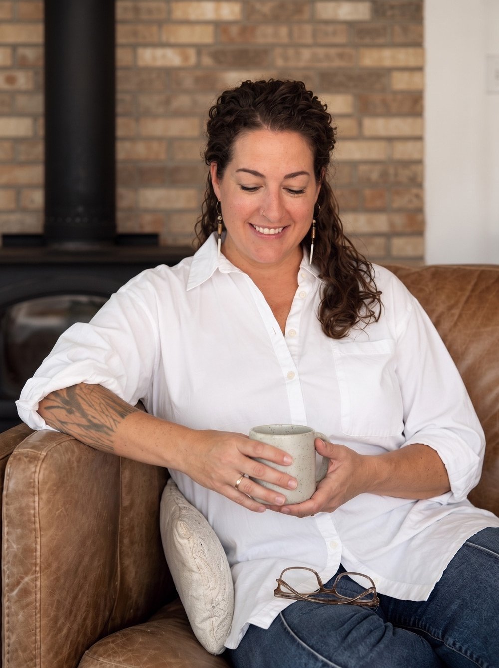 Grief, trauma, and relationship therapist Hannah Dudley is a woman with curly dark hair, wearing a white button-up shirt and hoop earrings, sitting on a tan leather sofa with a cushion, holding a ceramic mug and smiling.