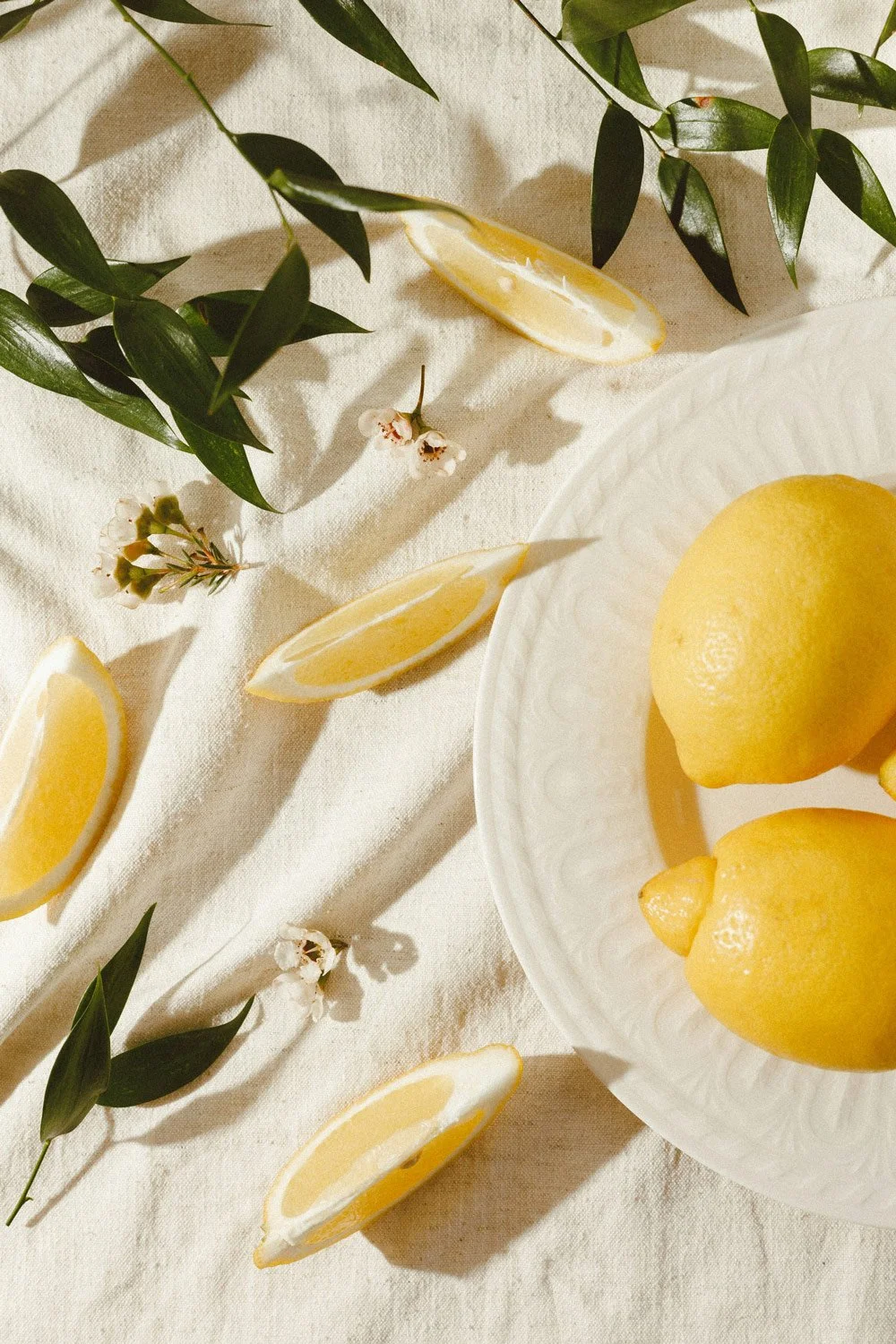 Lemons on a white plate and on a cream-colored tablecloth, with lemon wedges and green leaves scattered around.