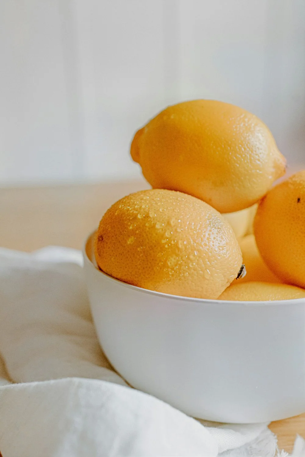 A white bowl filled with fresh yellow lemons on a light-colored surface with a white cloth underneath.