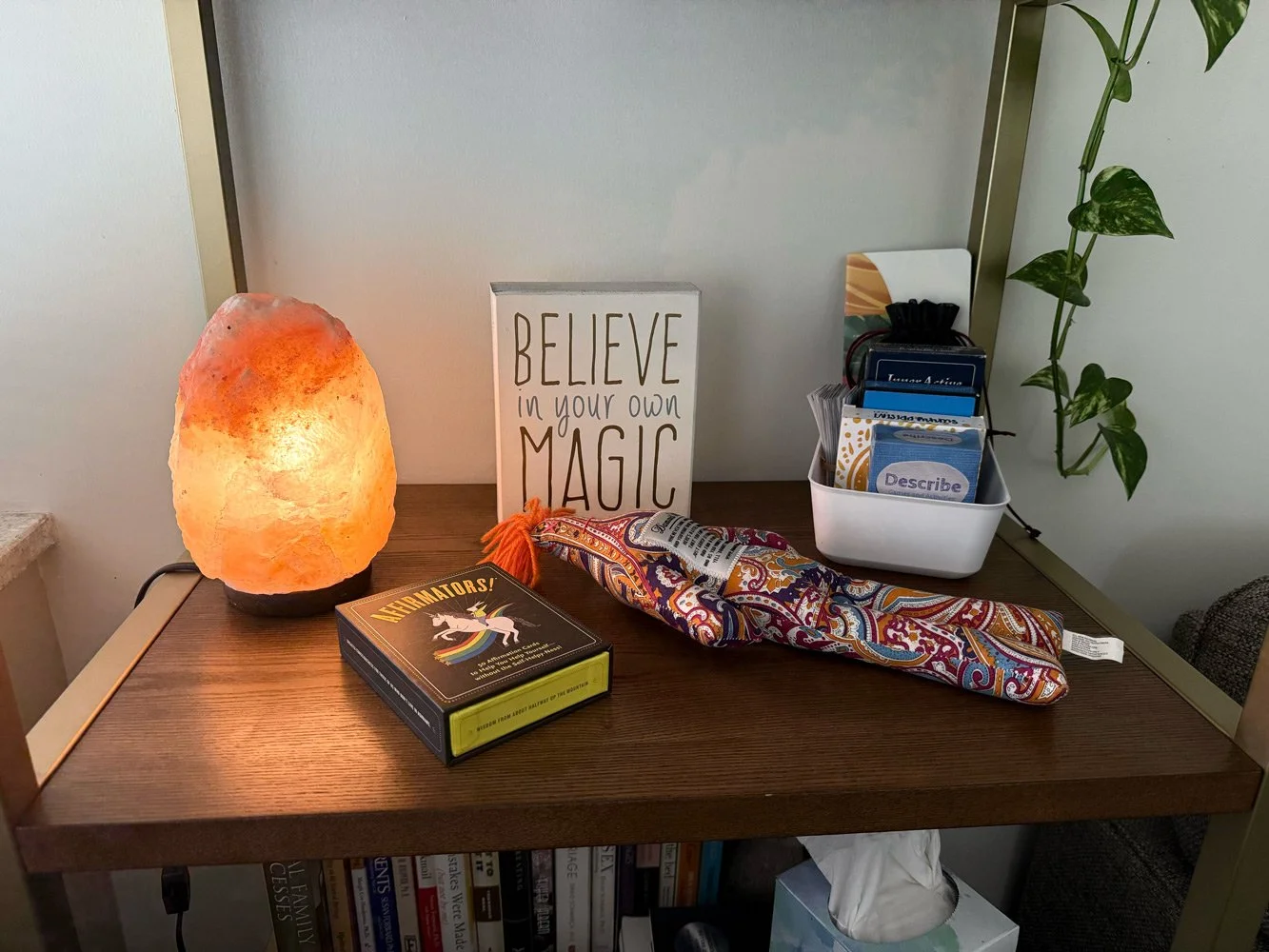 A wooden desk with a salt lamp on the left, a decorative sign saying 'Believe in your own magic' in the center, a colorful fabric item and a box of cards on top, and a white container holding various cards and papers on the right. There is a green pl