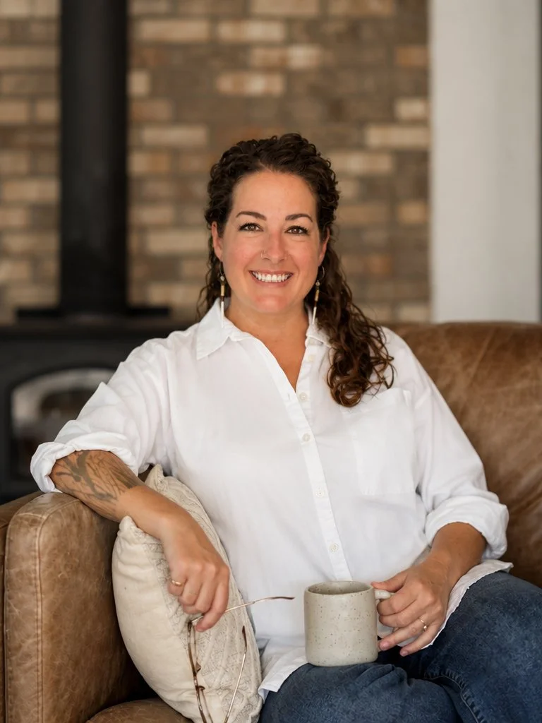 Hannah Dudley, LMFT, is a woman with curly brown hair, wearing a white shirt, smiles while sitting on a brown leather couch with a mug in her hand.