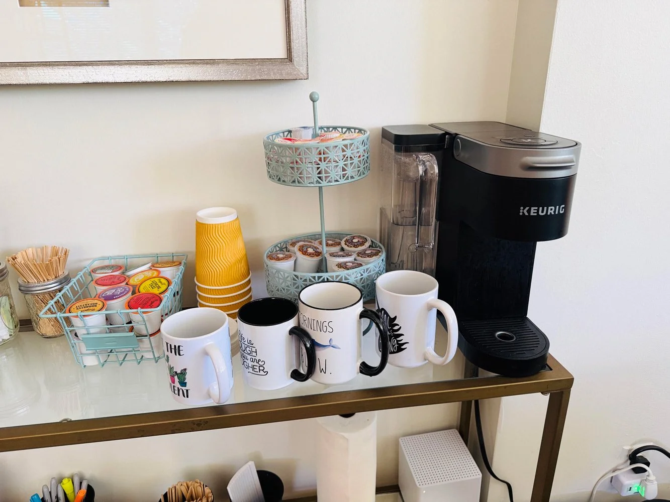 Coffee station with Keurig machine, mugs, coffee pods, disposable cups, and stirrers on a glass table.
