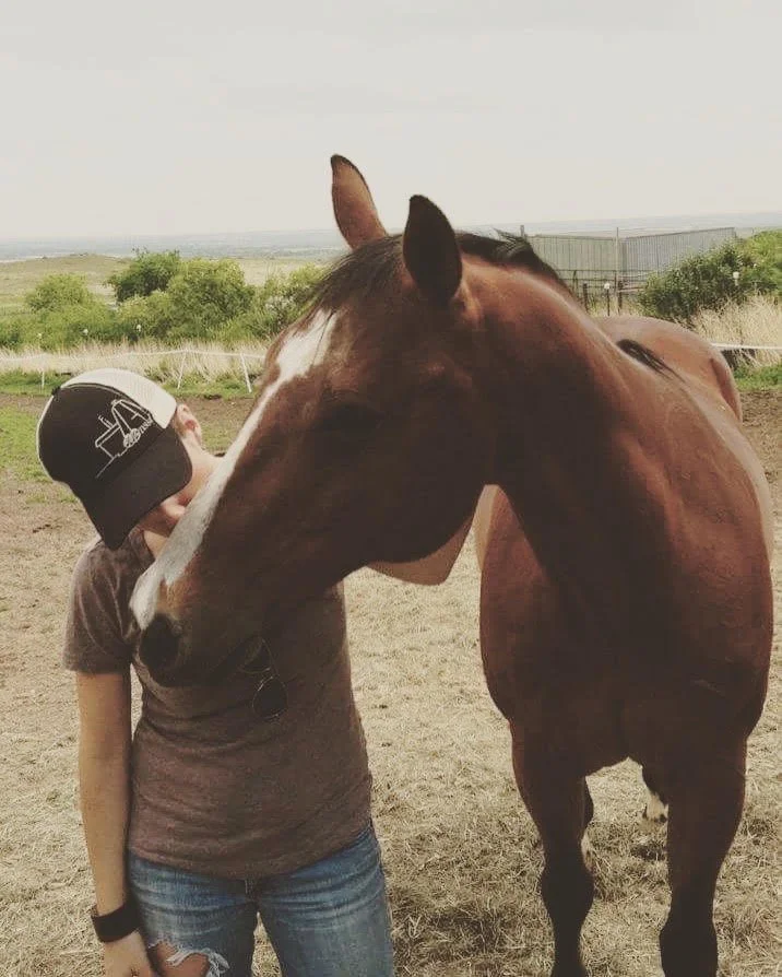 A vintage photo of grief, trauma, and relationship therapist Hannah Dudley as a girl with a baseball cap leaning her head against a brown horse's face in an open field.