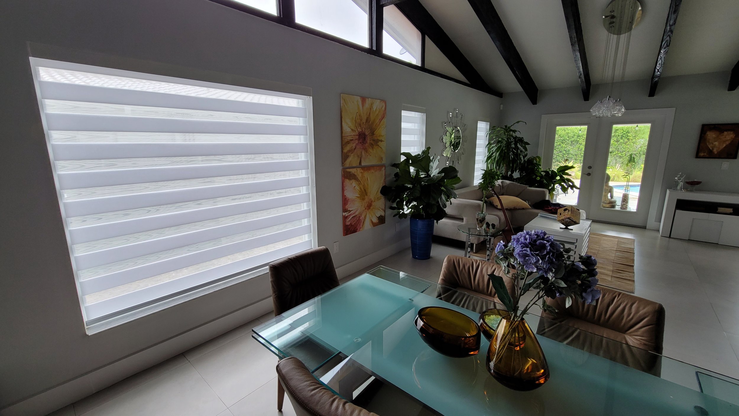 Living room with a large window with zebra blinds, colorful wall art, houseplants, a white sofa, a glass coffee table with decorative vases, and a pool view outside through glass door.