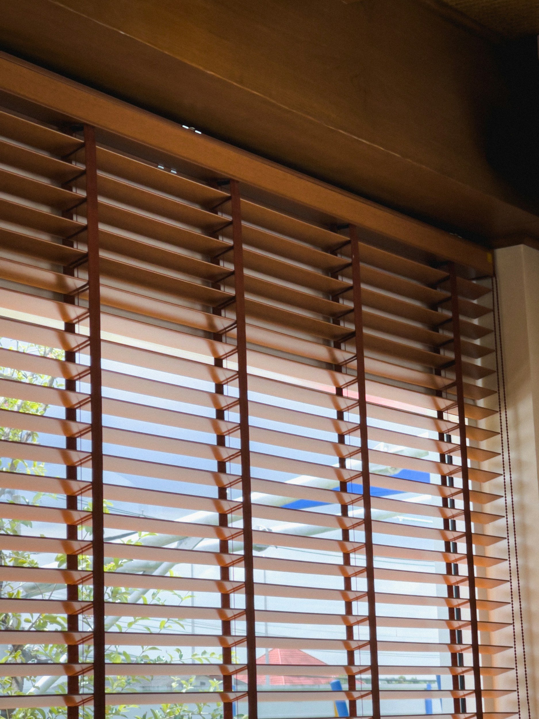 View of a window with wooden horizontal blinds partially open, revealing a bright daytime sky and some outdoor structures and greenery.