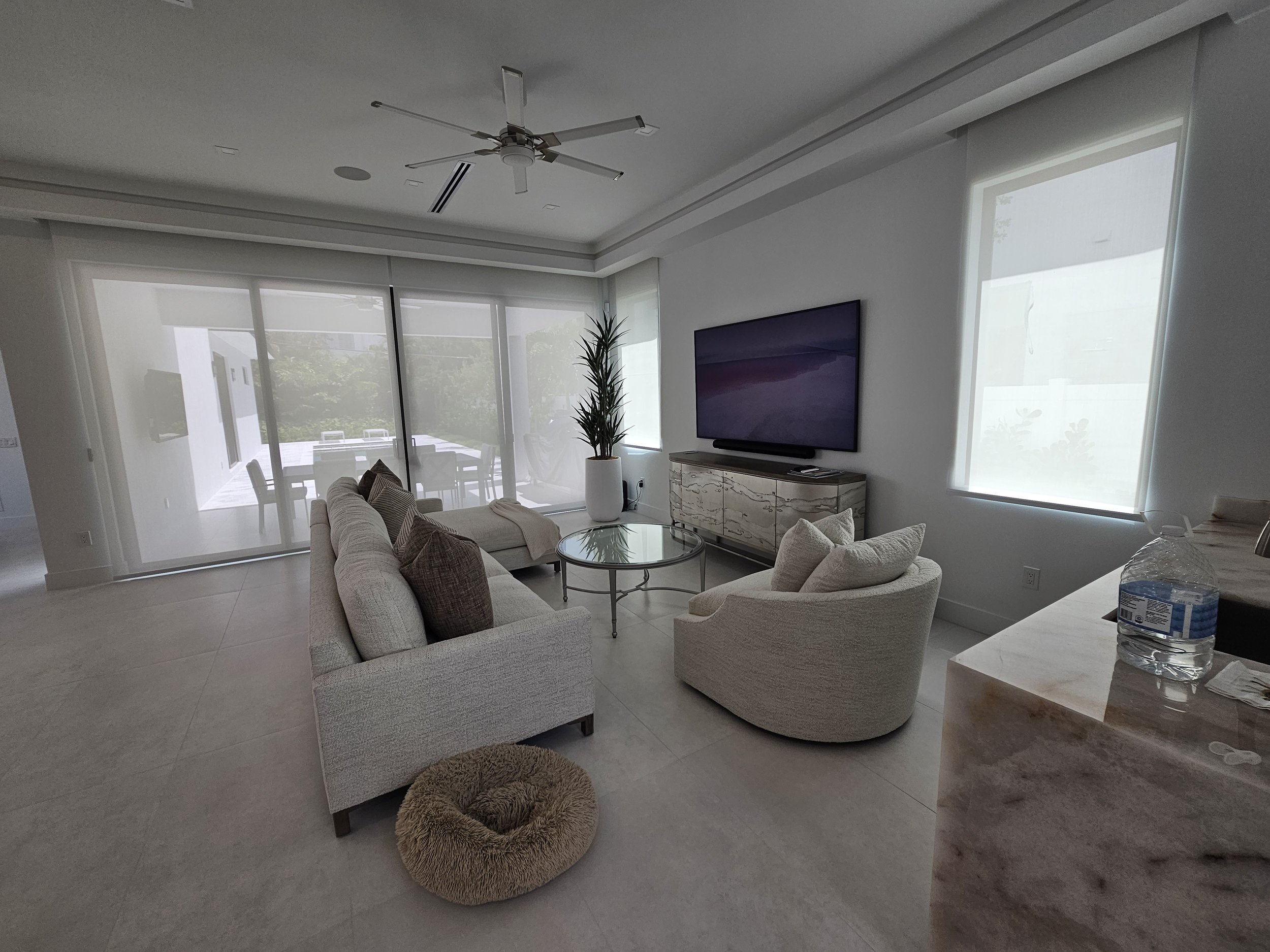 Living room with beige sofas, a potted plant, a large flat-screen TV on a cabinet, and windows with blinds letting in natural light.
