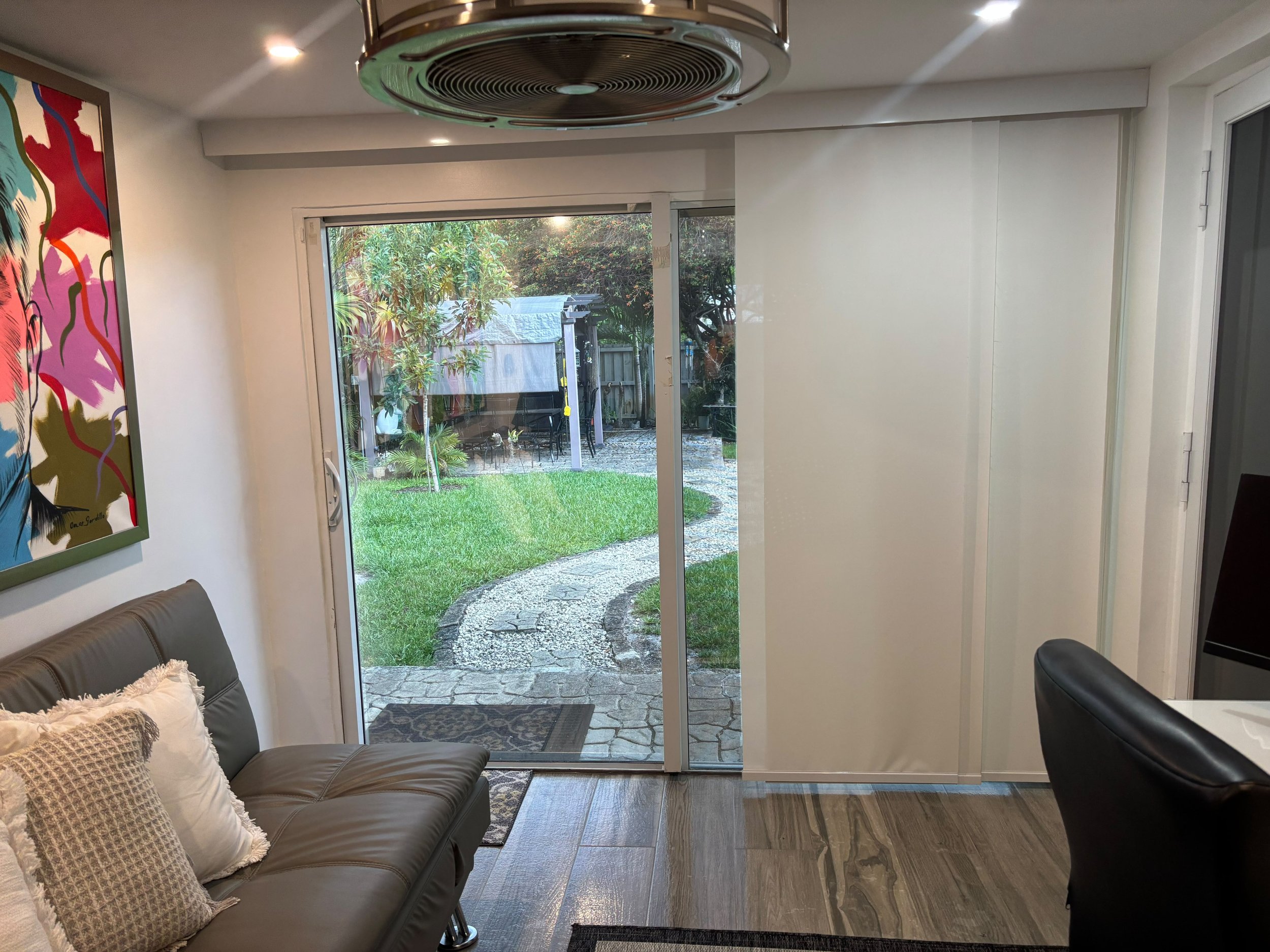 Image shows an interior room with a sliding glass door leading outside to a backyard with green grass, trees, and outdoor furniture. Part of a gray leather couch with pillows is visible, and a colorful abstract painting is on the wall to the left.