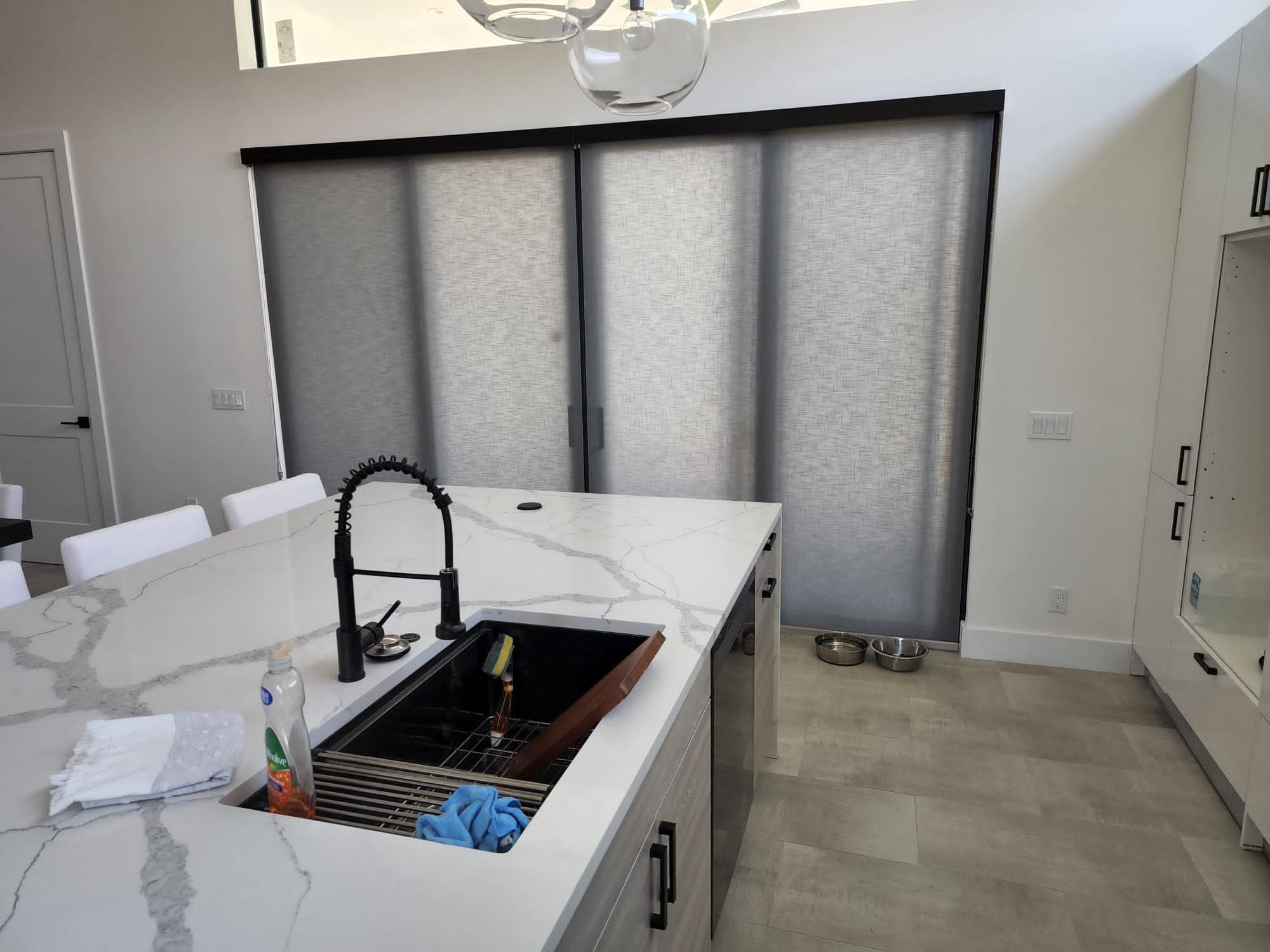 A modern kitchen with a white marble island, a black pull-down faucet, and a gray backsplash. There are white chairs around the island, with dish soap and cleaning cloth present. A pair of dog bowls is on the floor next to a white cabinet, and metal pet bowls on the floor.