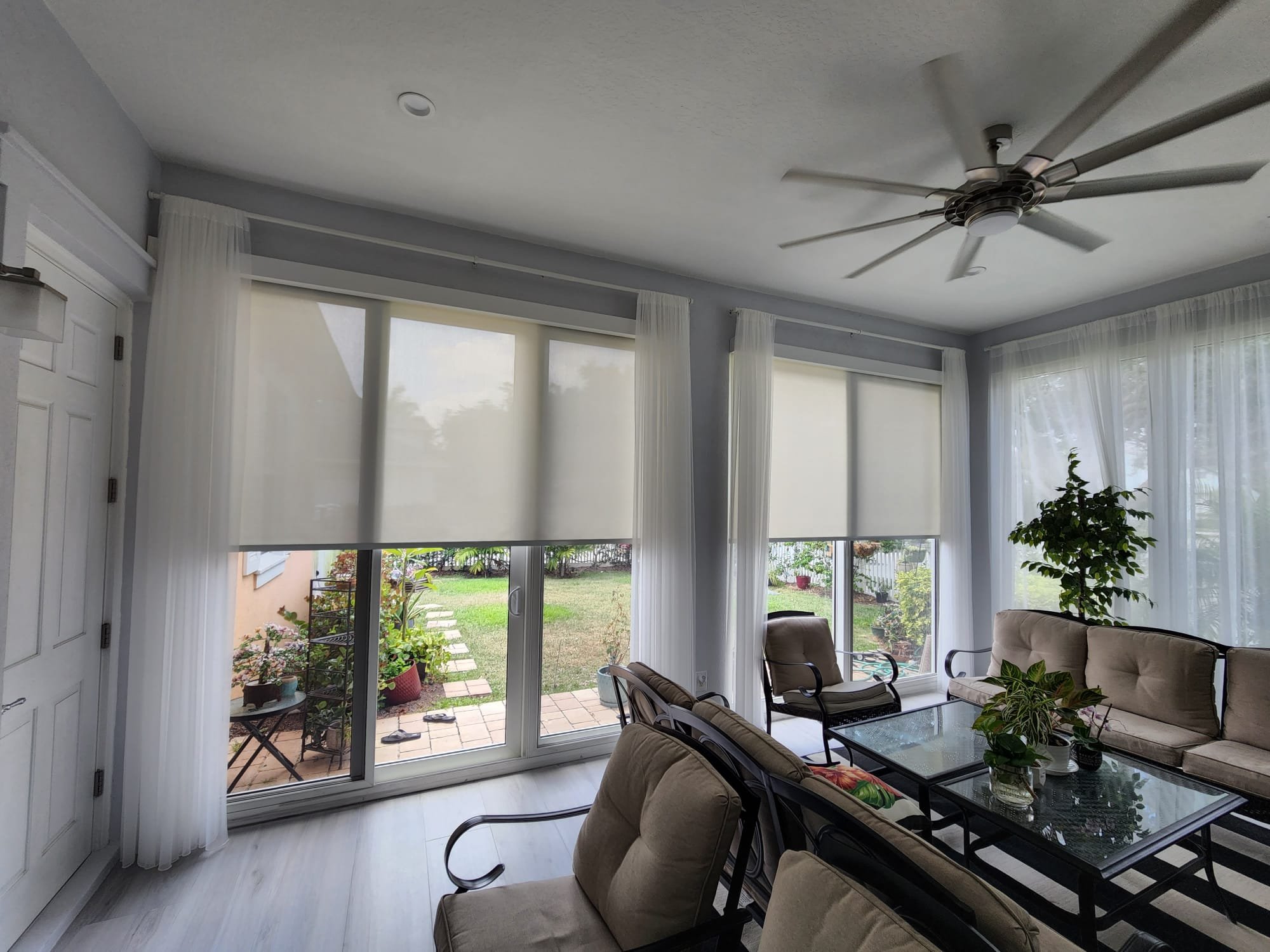 Living room with large windows and sliding glass doors, white curtains, ceiling fan, beige couches, glass coffee table with decorative plants, and outdoor patio with garden plants visible through the windows.