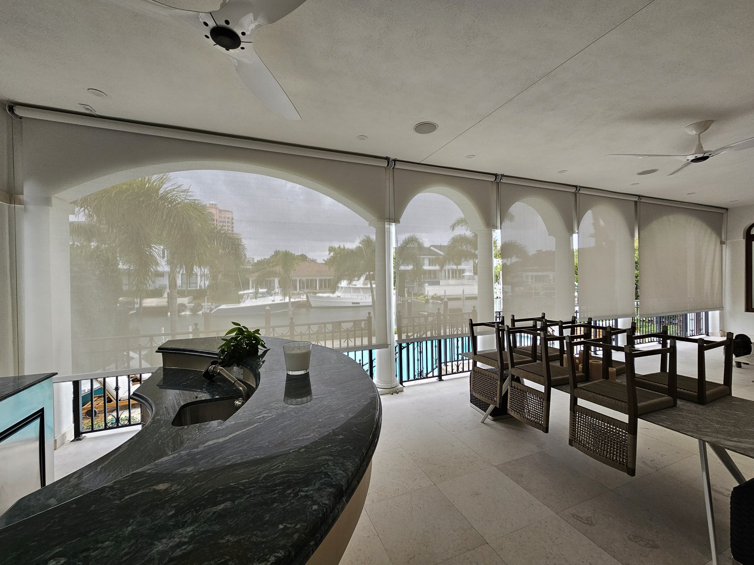 A covered outdoor patio area with ceiling fans, large windows with roller shades, palm trees and boats visible outside, and seating including chairs and a table.