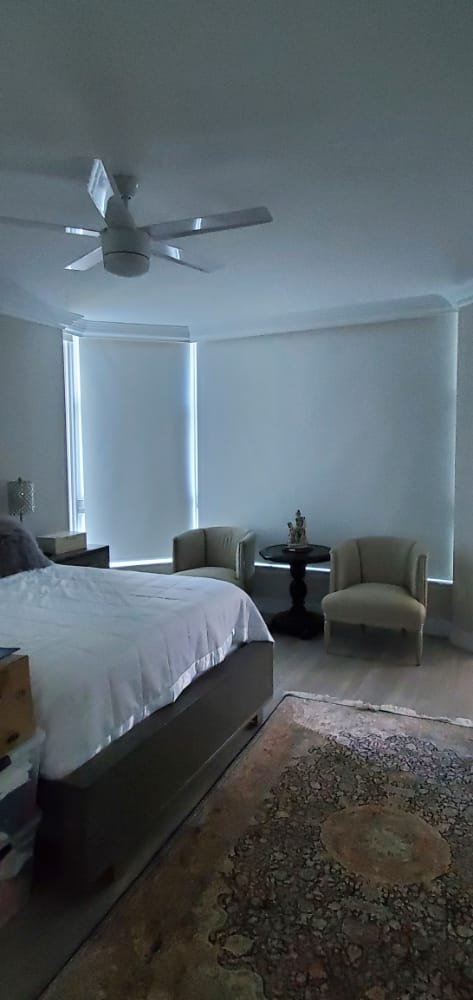 A bedroom with a bed, two beige chairs, and a small black round table, with blinds covering the windows and a patterned rug on the floor.