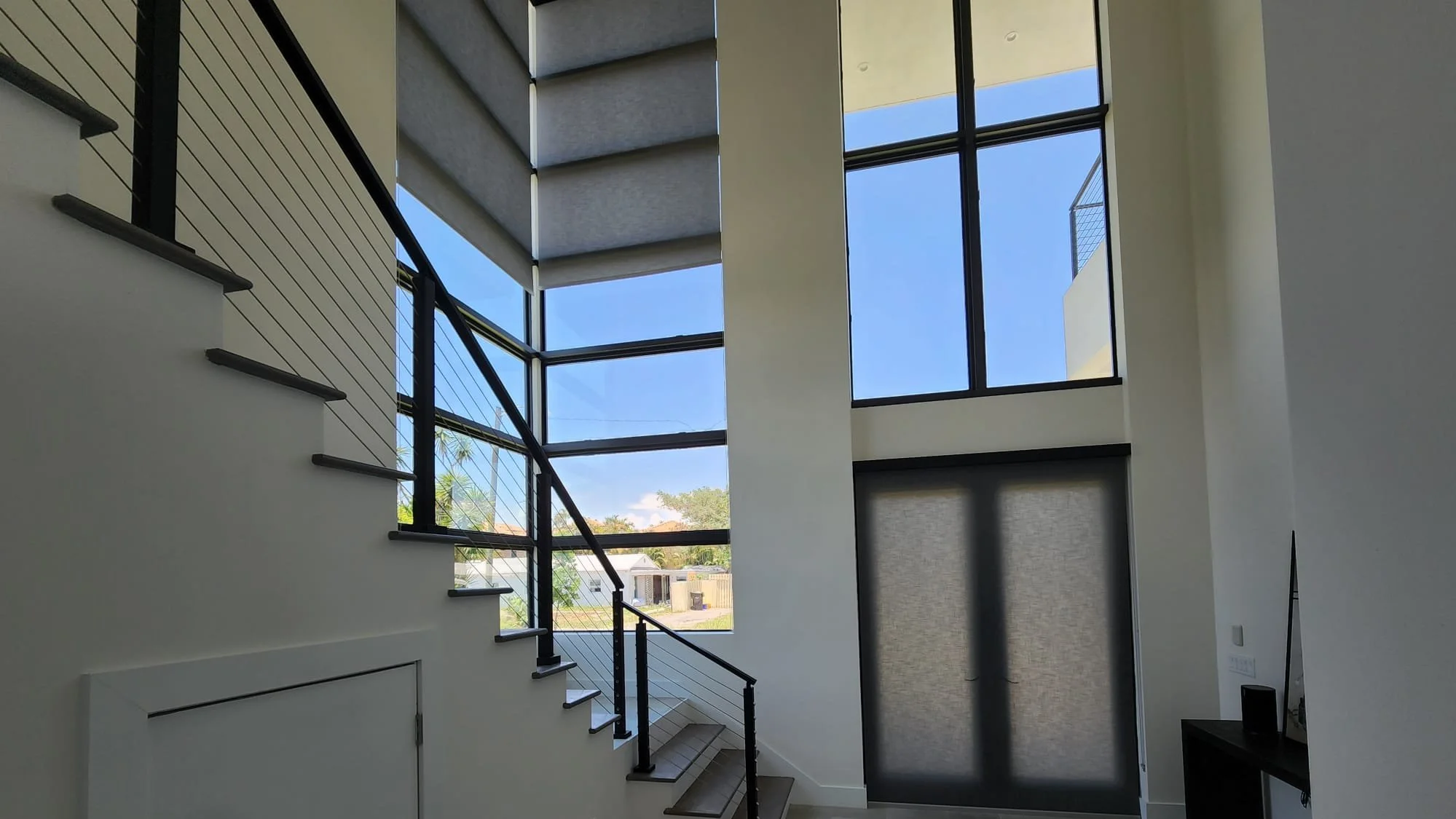 Interior view of a modern home with large stairs, black handrail, large windows with blinds, and a view of the neighborhood outside under a bright blue sky.