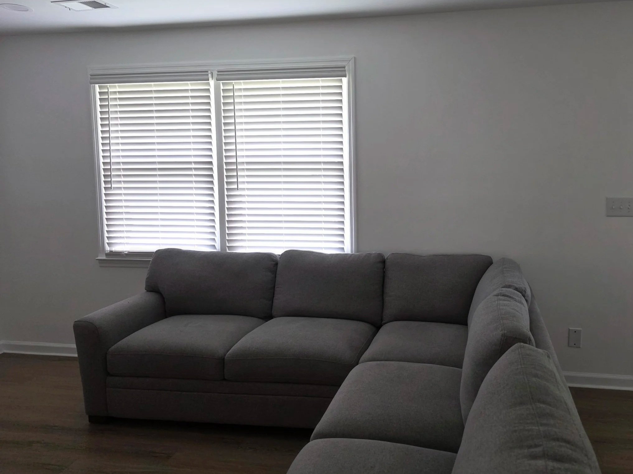 A living room with a gray sectional sofa in front of a window with closed white blinds. The walls are painted light gray, and there are electrical outlets on the right side of the image.