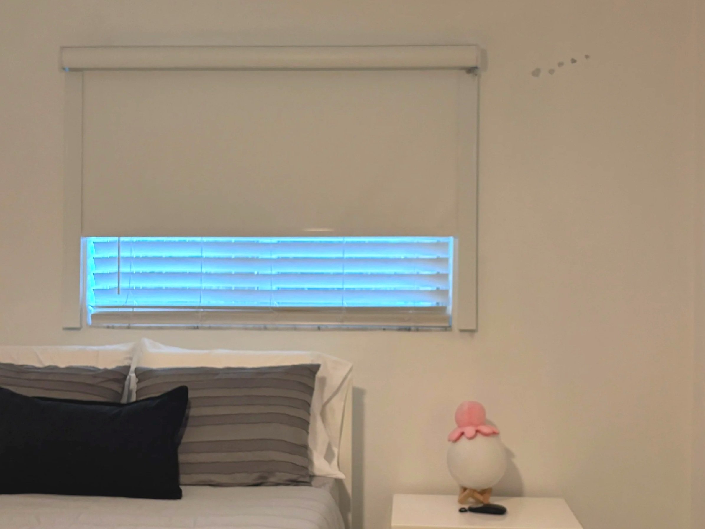 A bedroom with dual window treatments, a blackout roller shade with side channels and horizontal faux wood blinds for light control.