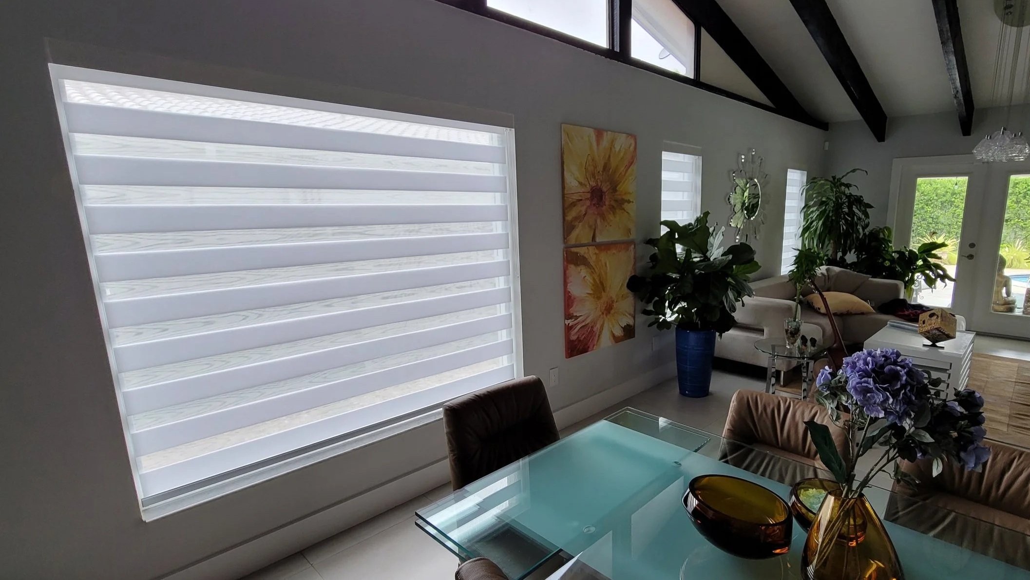 Living room with large windows, white walls, potted plants, wall art, beige sofa, glass-top table, and flowers in a vase.
