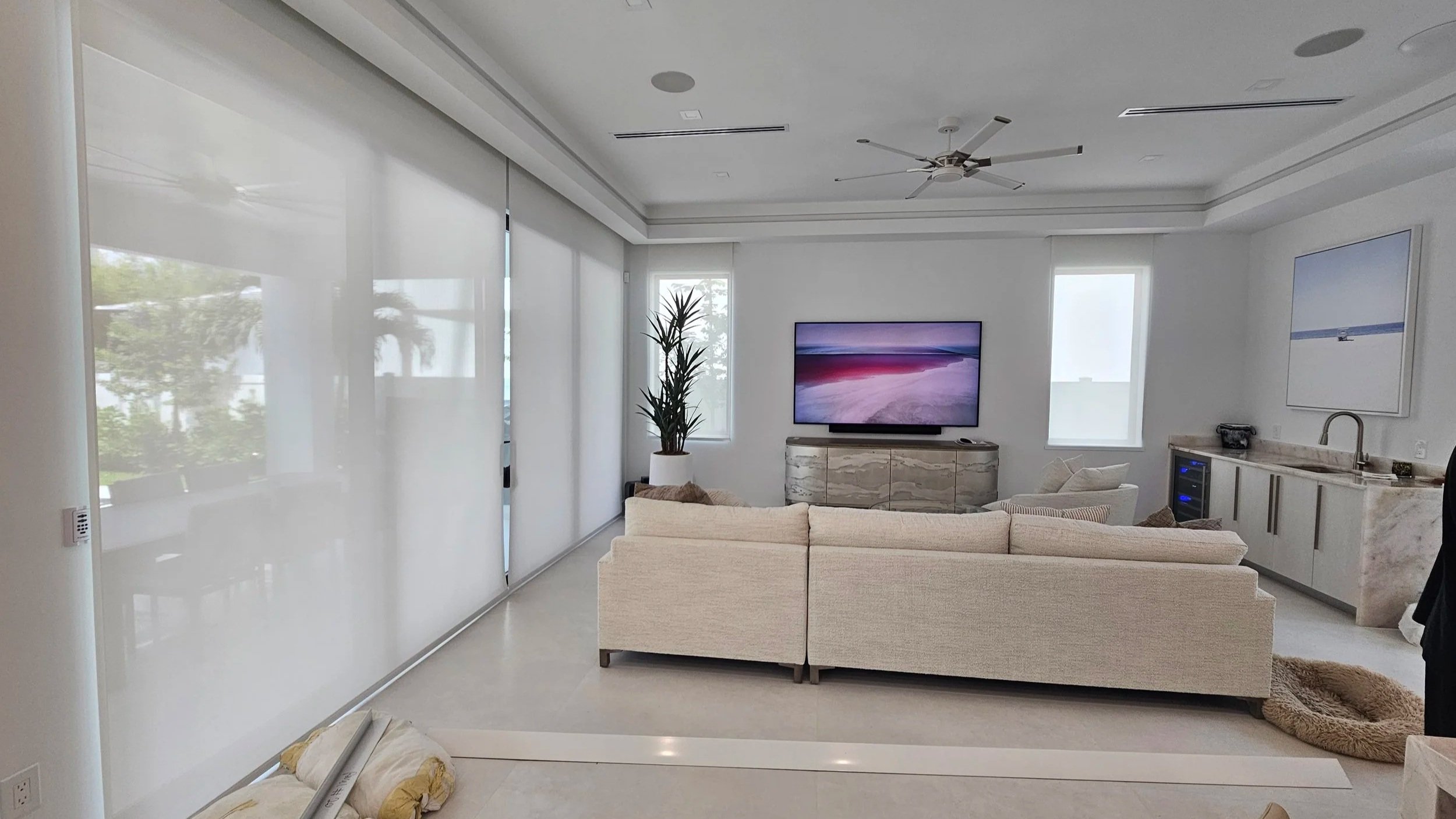 Modern living room with white walls, large windows with blinds, a ceiling fan, a beige sofa, a TV on a marble cabinet, a tall plant, and a kitchen area with a marble countertop and appliances.