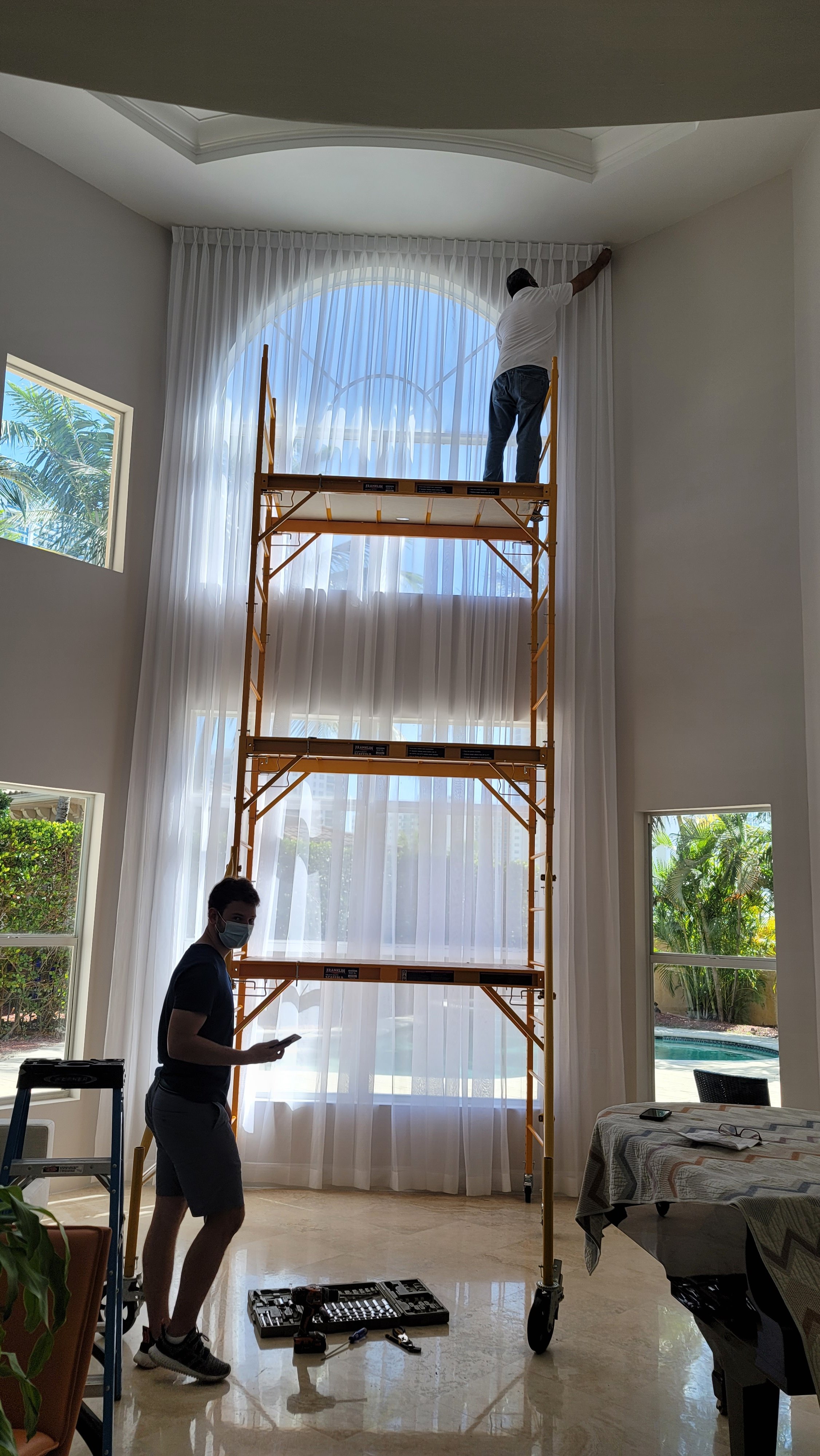 Two people installing or repairing high pleated curtains in a bright living room with large, arched windows and tropical view outside.