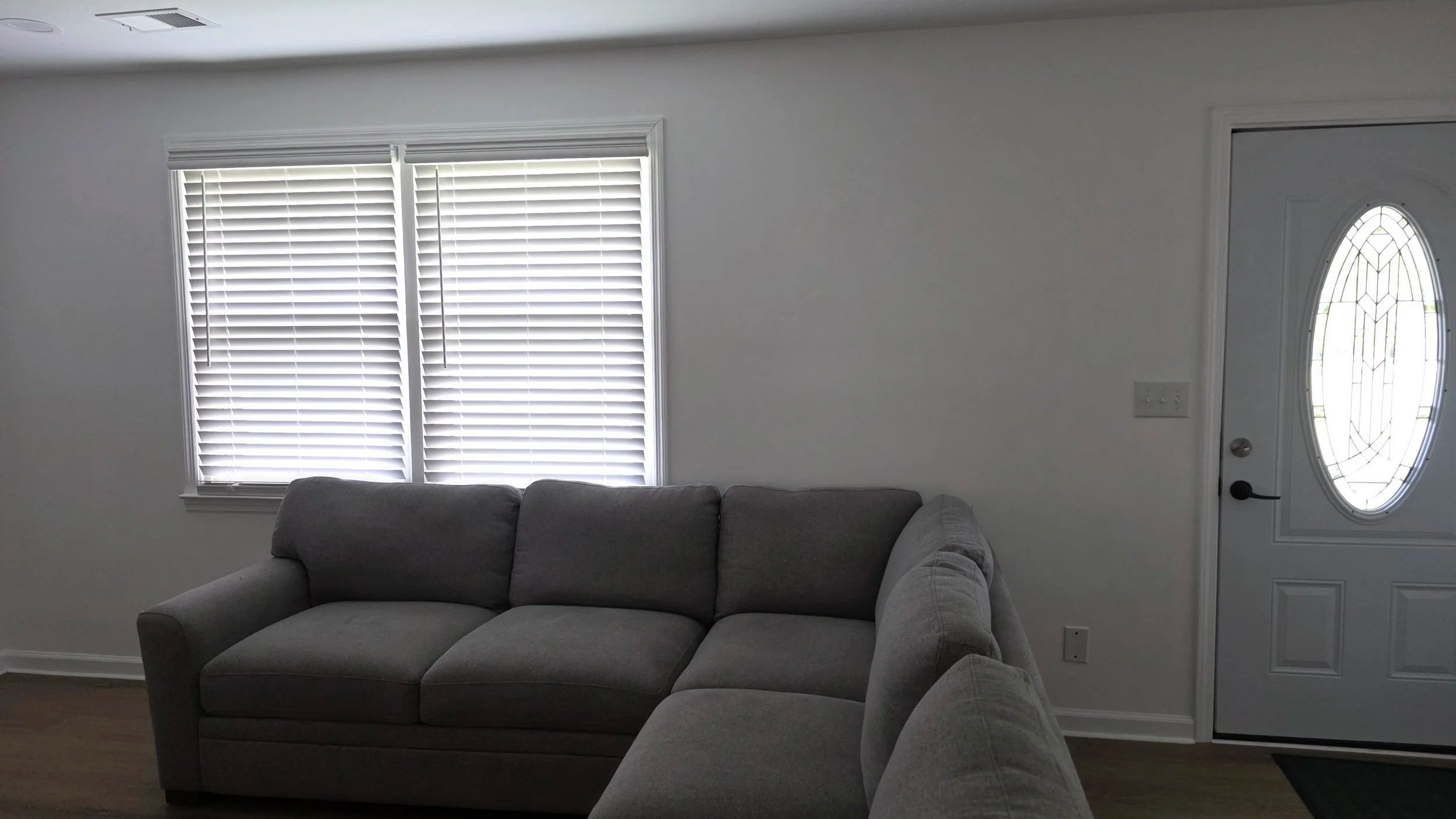 Living room corner with a gray couch and a large window with white blinds, next to a white front door with an oval glass panel.