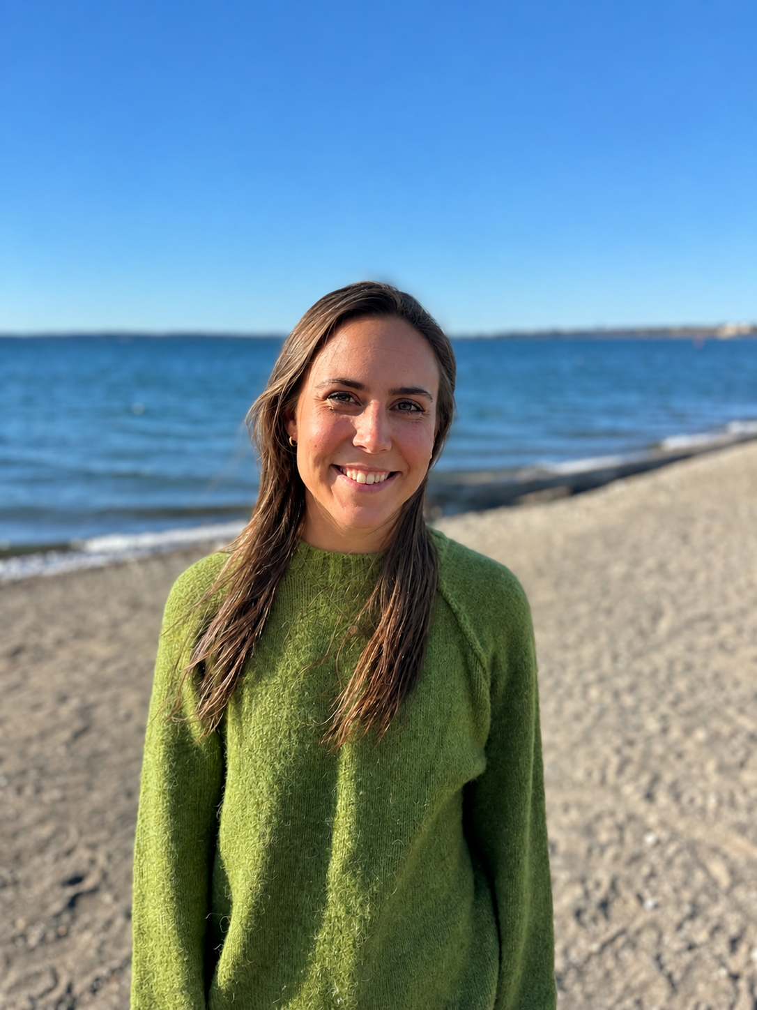 A woman with long brown hair smiling at the camera on a sandy beach with the ocean and blue sky in the background, wearing a green sweater.