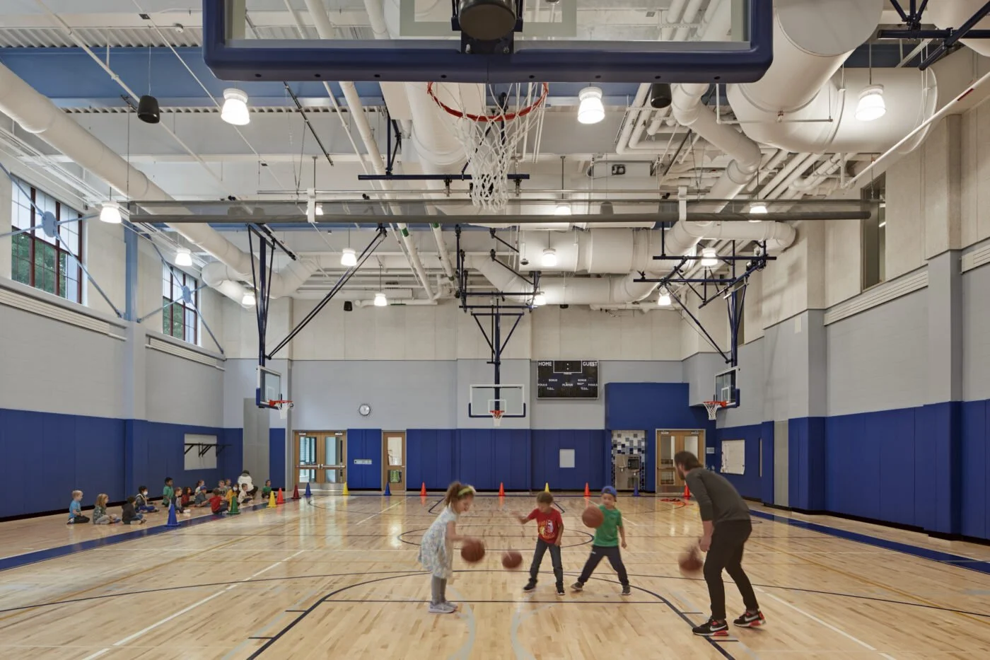 A group of children practicing basketball under the supervision of an adult coach in a spacious indoor gymnasium.