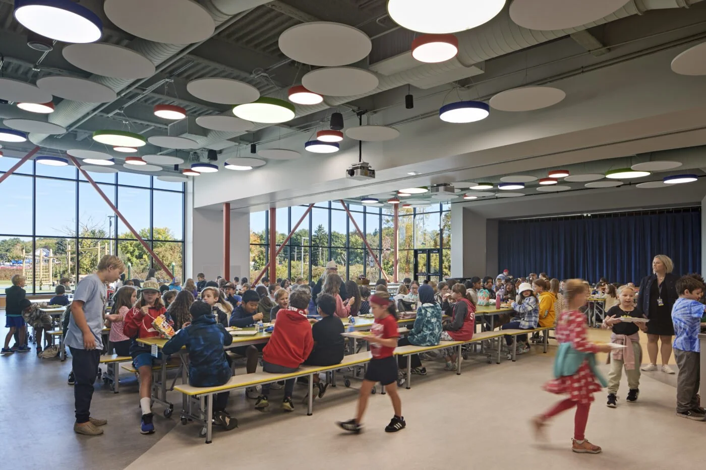 Children sitting at tables in a spacious, brightly lit cafeteria or multipurpose room with large windows and colorful ceiling lights.