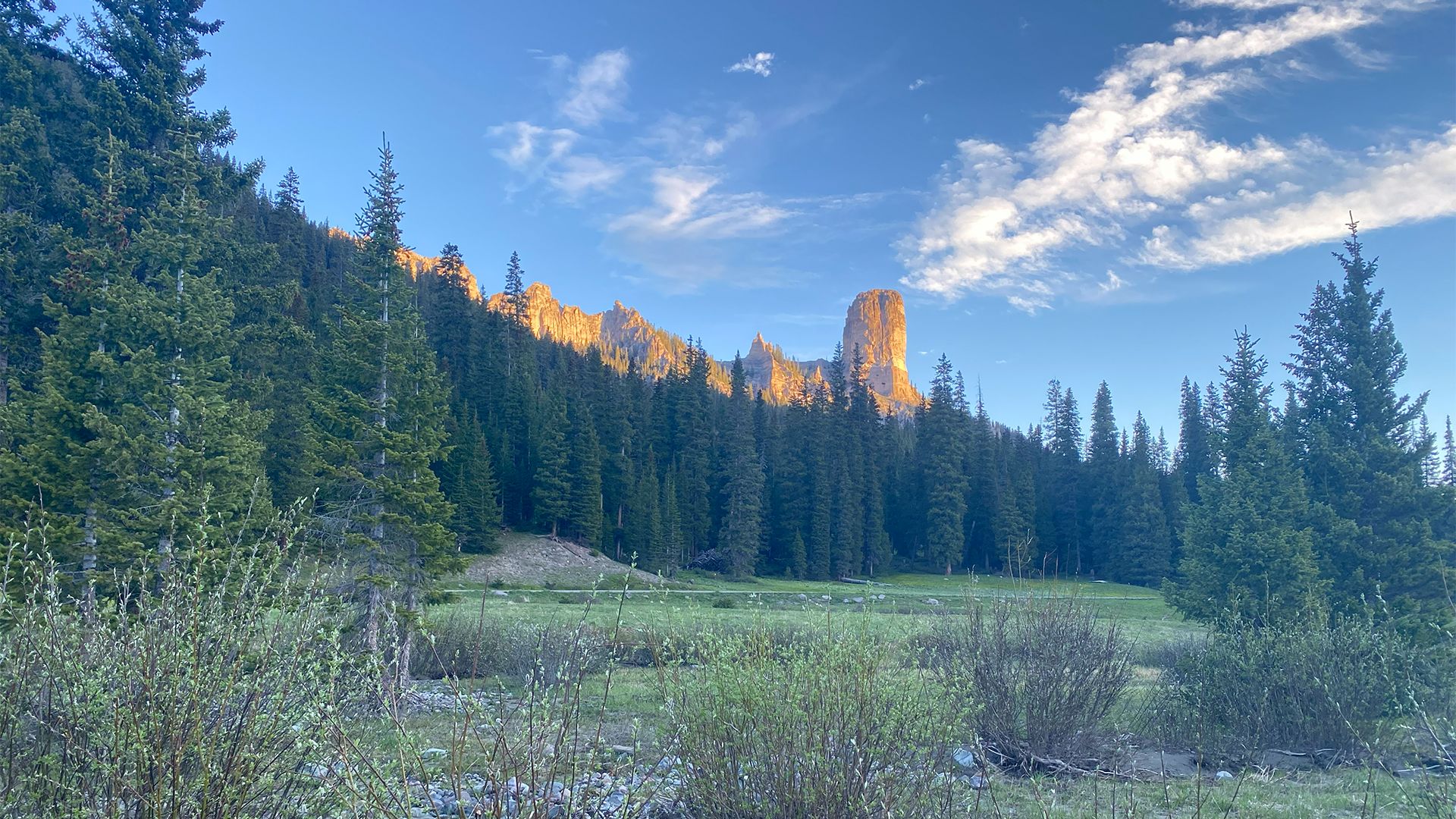Sunlit granite rock formations rising above a dense evergreen forest and meadow beneath a blue, cloud-streaked sky