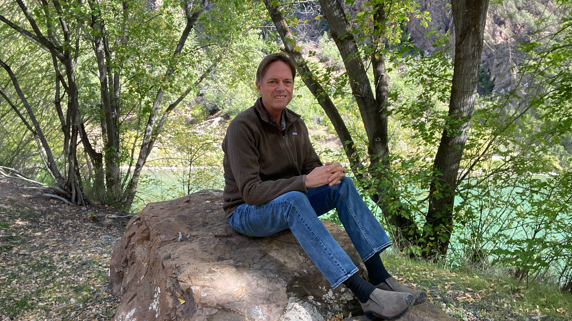 Man sitting on a large rock beside a turquoise river in a wooded canyon